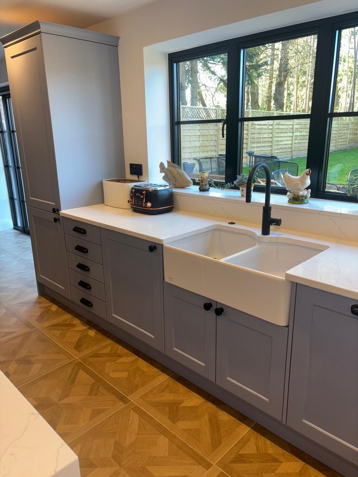 A kitchen featuring grey cabinets, a white farmhouse sink, black faucet, and light countertops with herringbone flooring.