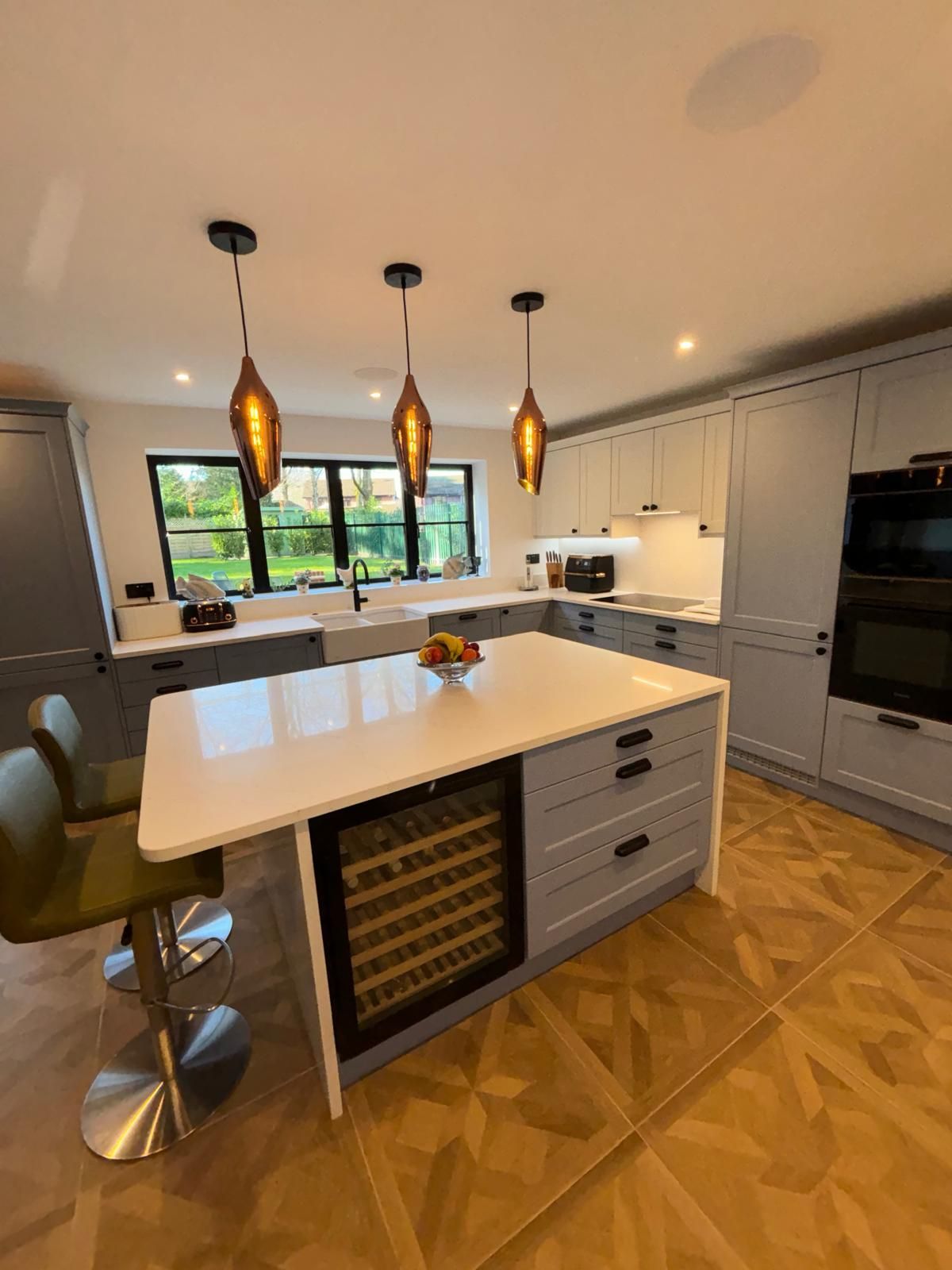 A modern kitchen with a large white island, grey cabinets, a wine fridge, three bronze pendant lights, and wood flooring.