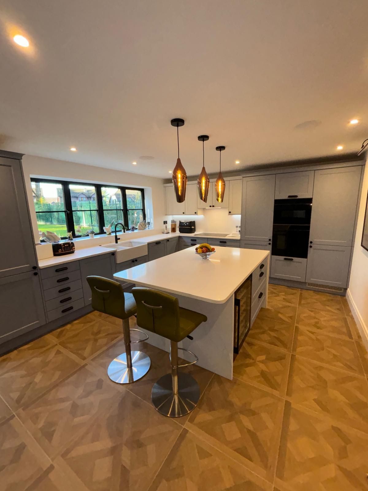 Modern kitchen with a white island, two olive green stools, grey cabinetry, and three hanging lights over wooden floors.