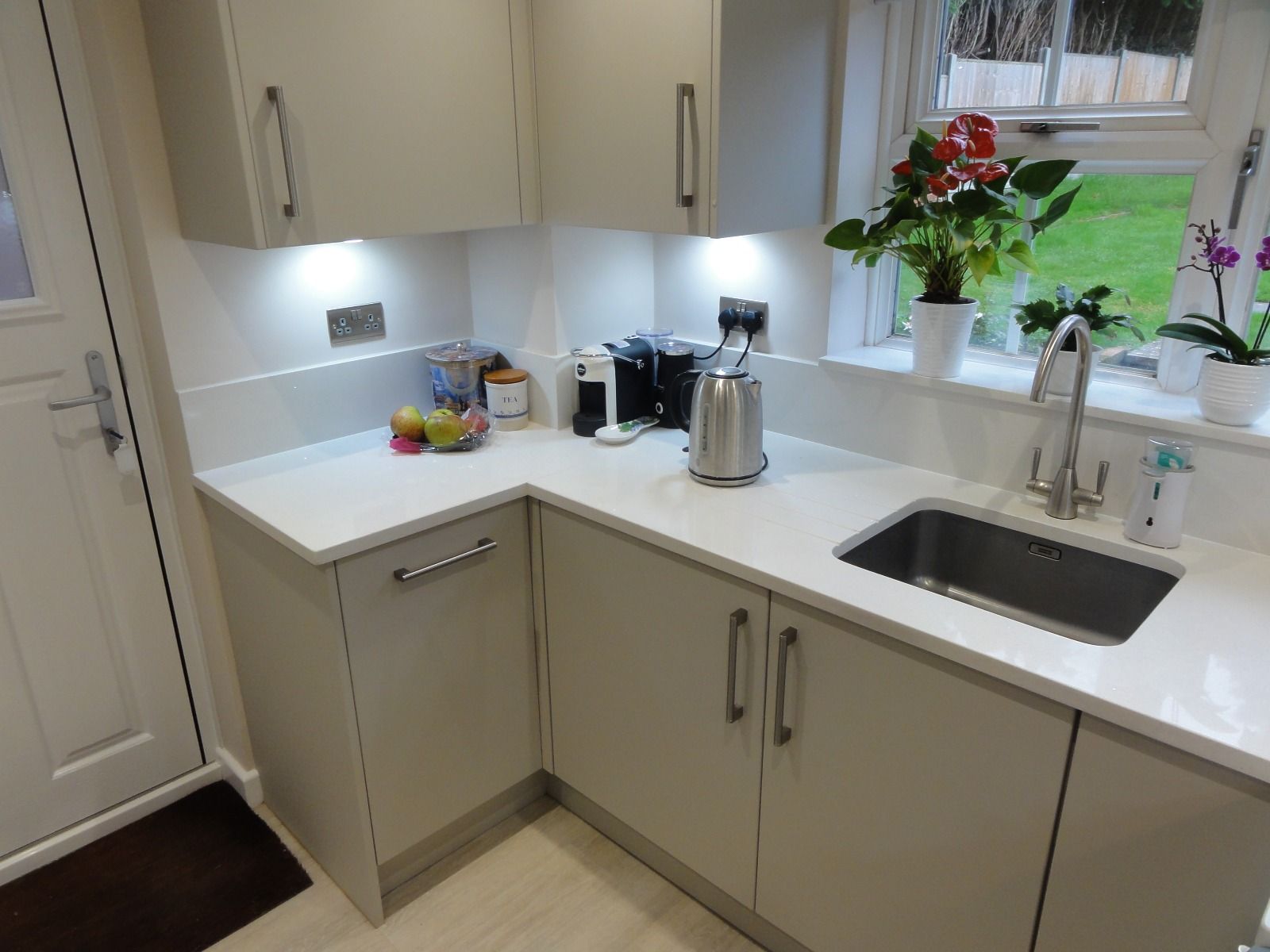 A light grey kitchen corner with white countertops, a sink, a coffee station, and potted plants on a window sill.