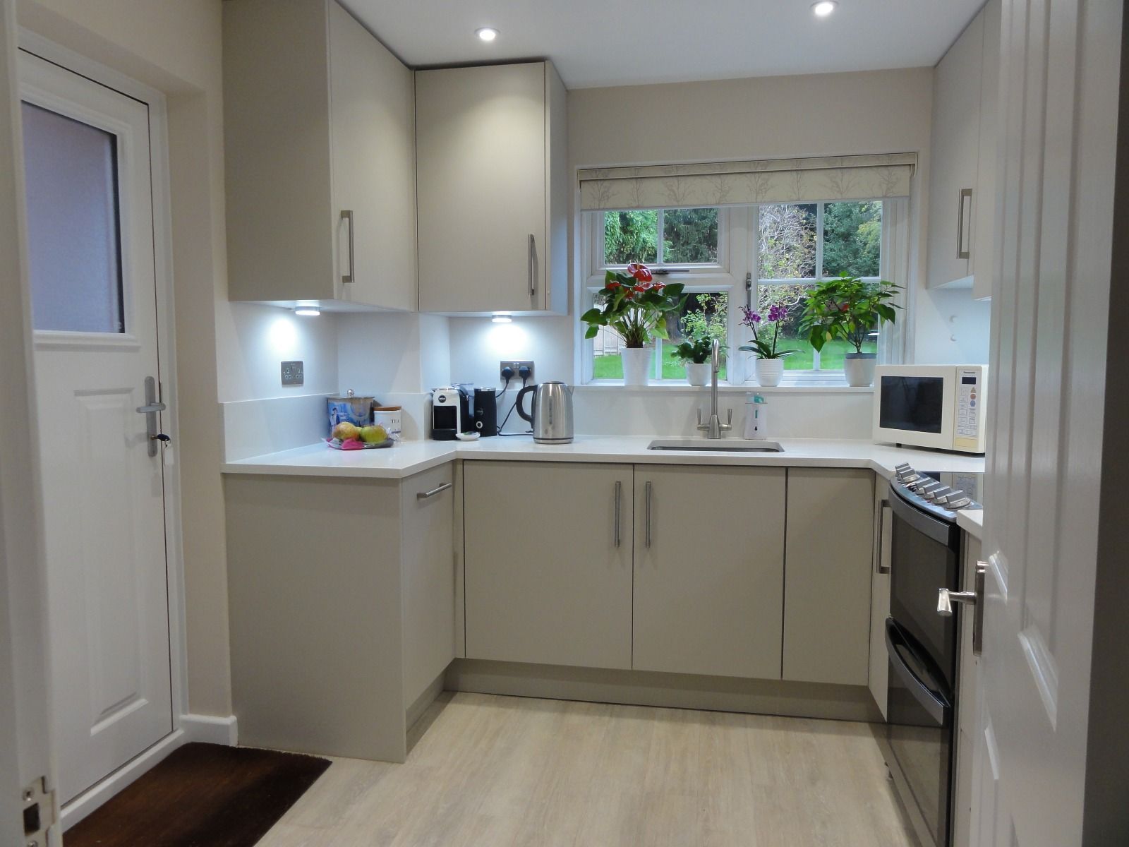 A modern, neutral-toned kitchen with light cabinetry, white countertops, a sink by a window, and stainless steel appliances.