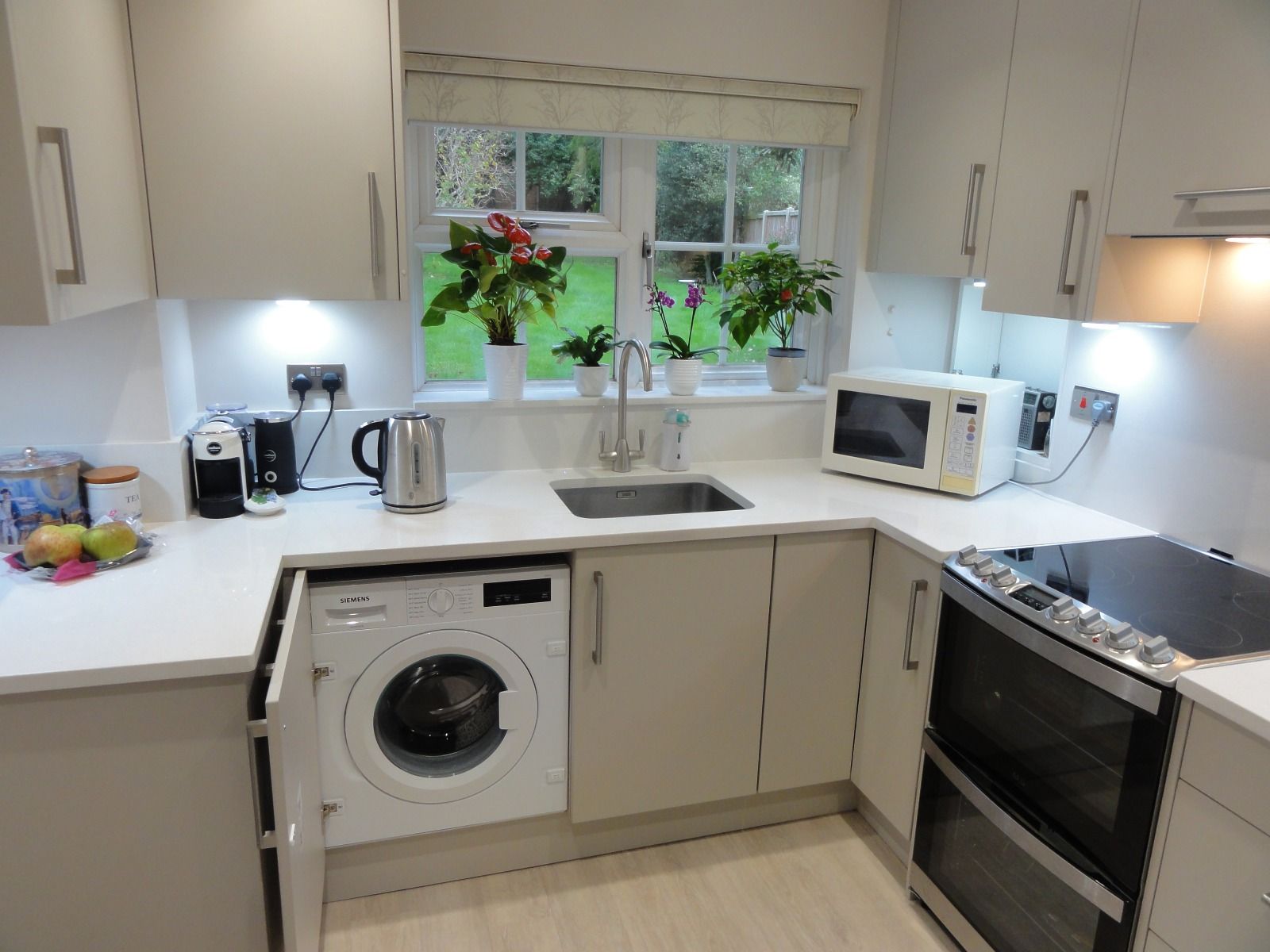 A modern kitchen with light grey cabinets, white countertops, a sink, a built-in washing machine, and an electric stove.