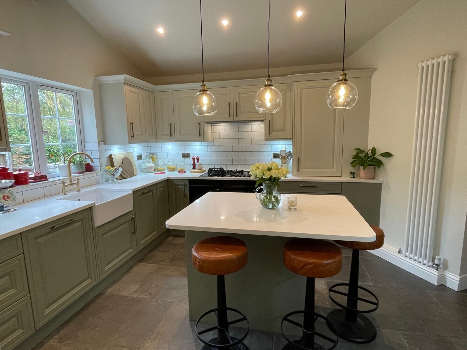 A bright, modern kitchen featuring sage green cabinets, a white island with three stools, and three globe pendant lights.