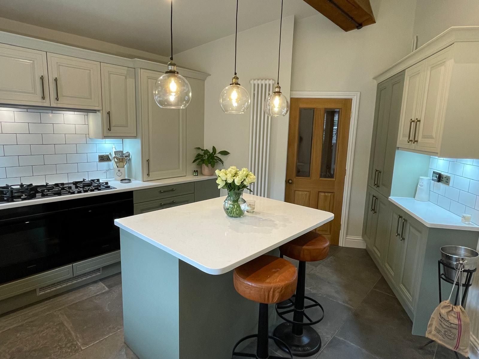 A sage green kitchen with a white-topped island, bar stools, three pendant lights, and stainless-steel appliances.