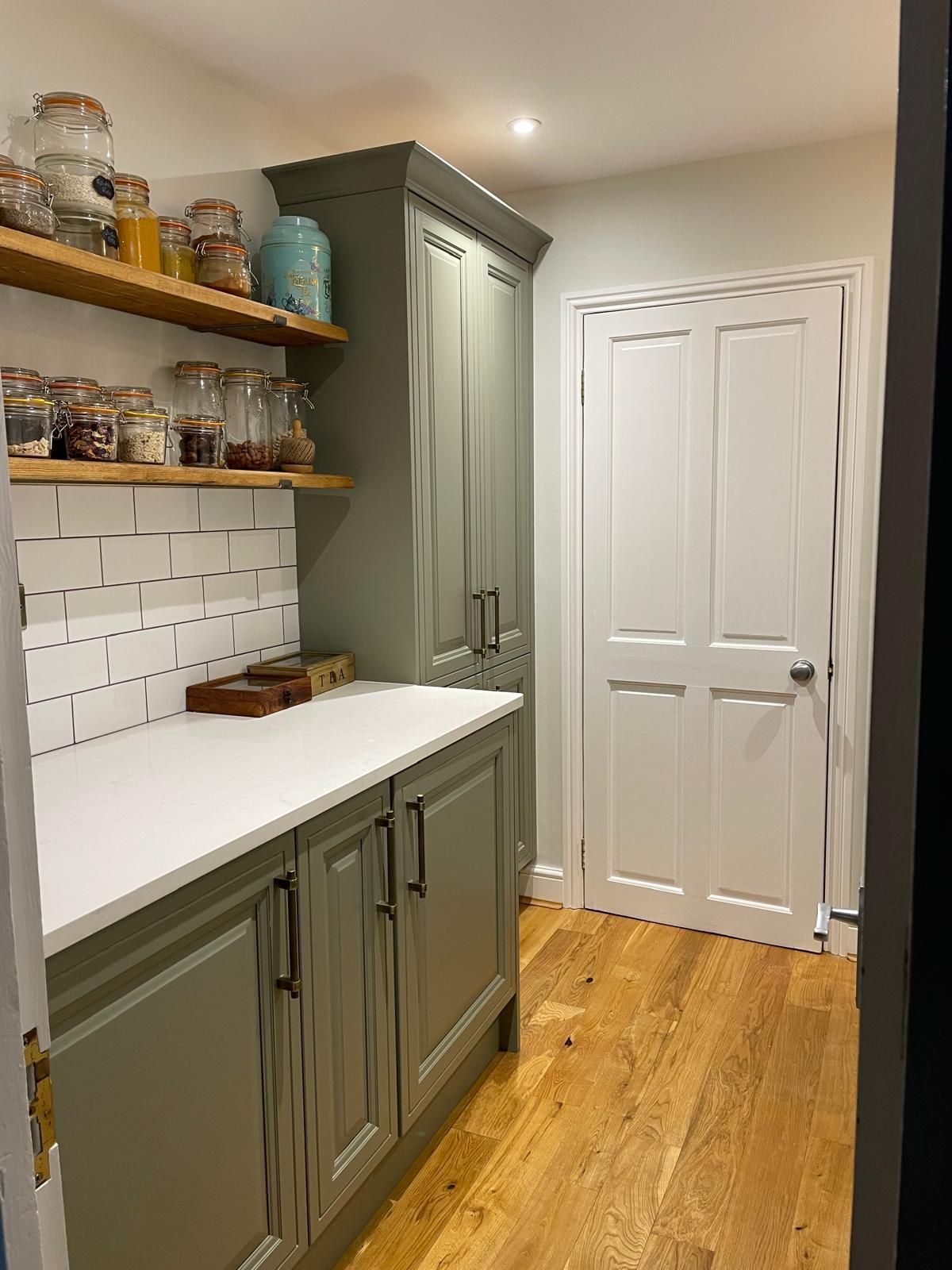 A pantry with sage green cabinets, white countertops, a subway tile backsplash, open wood shelving, and a white door.