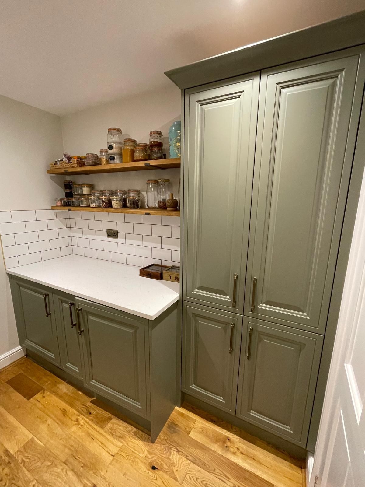 A kitchen featuring sage green cabinets, a white countertop, wooden floating shelves with glass jars, and a tiled wall.