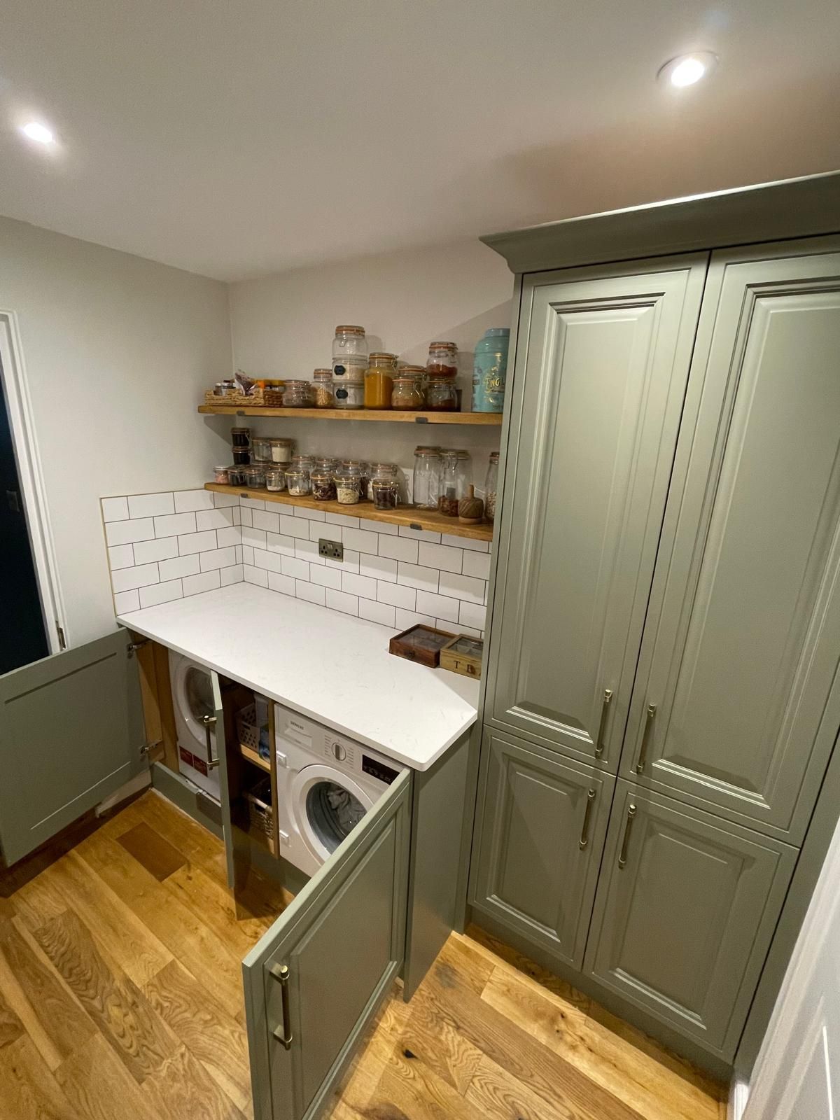 A laundry room with sage green cabinets, wood floors, a white counter over two machines, and open wood shelving.