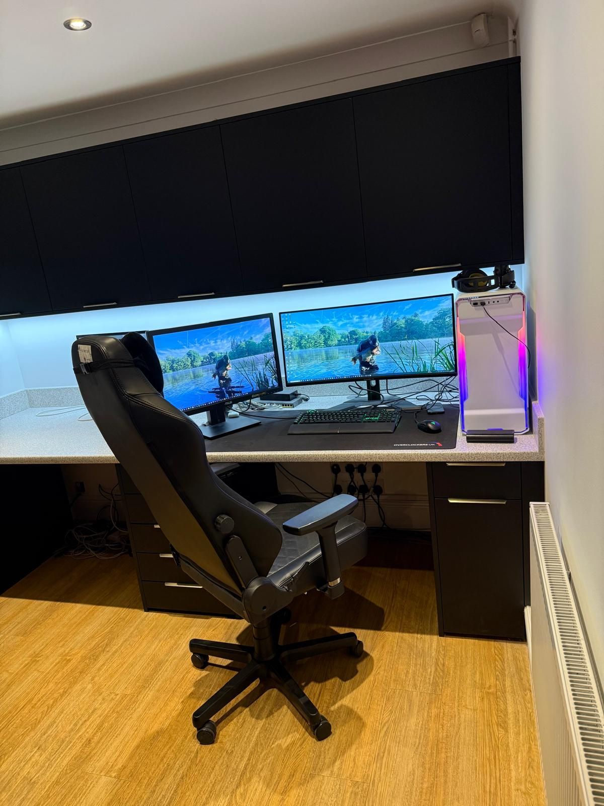 A dual-monitor gaming setup with a black ergonomic chair on a wooden floor in a home office.