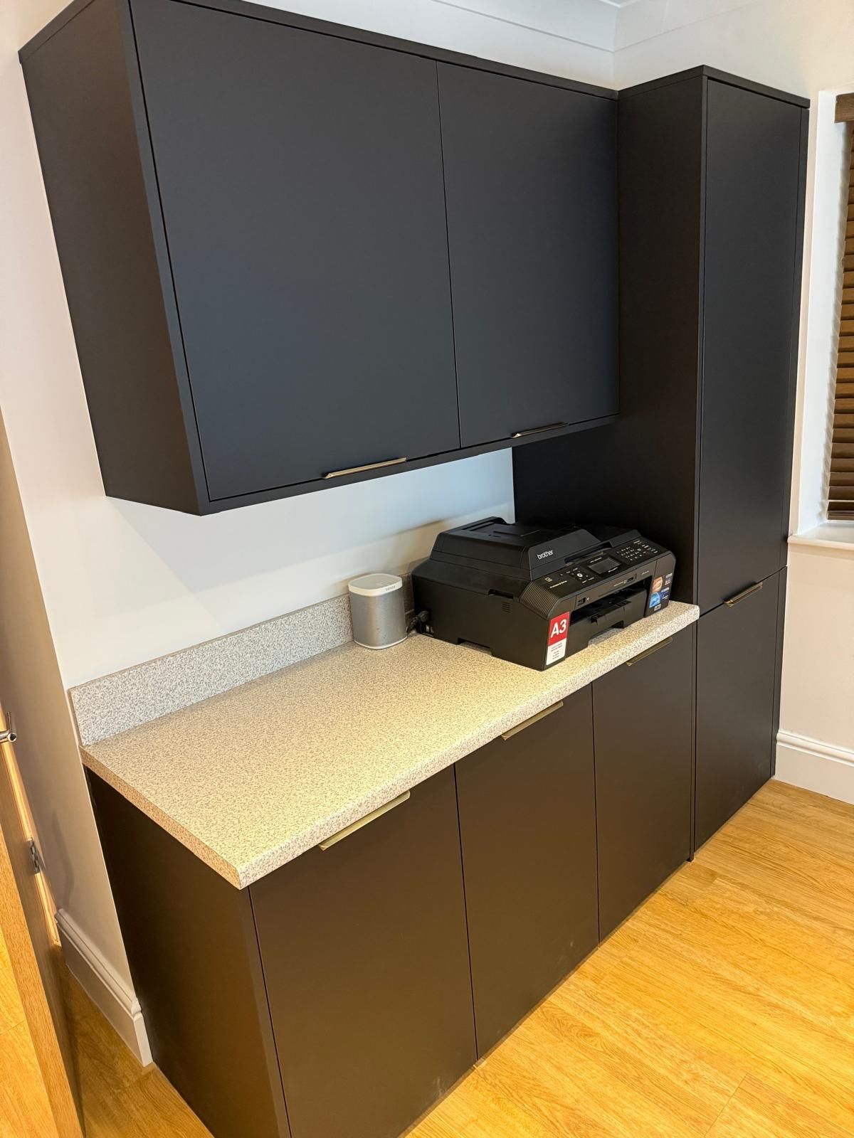 A home office setup featuring matte black cabinets, a light speckled countertop, a printer, and a smart speaker.