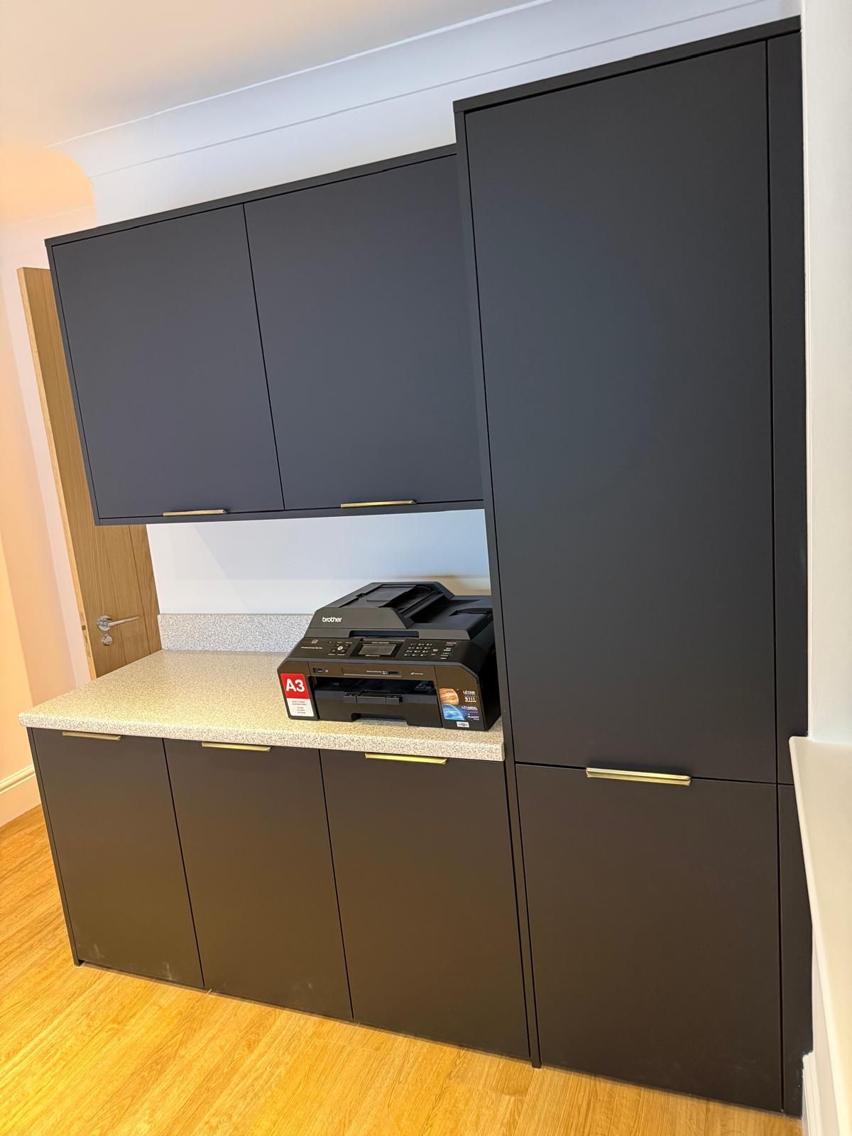 Matte black kitchen cabinetry with gold handles, a speckled countertop, and a black printer, set on a light wood floor.