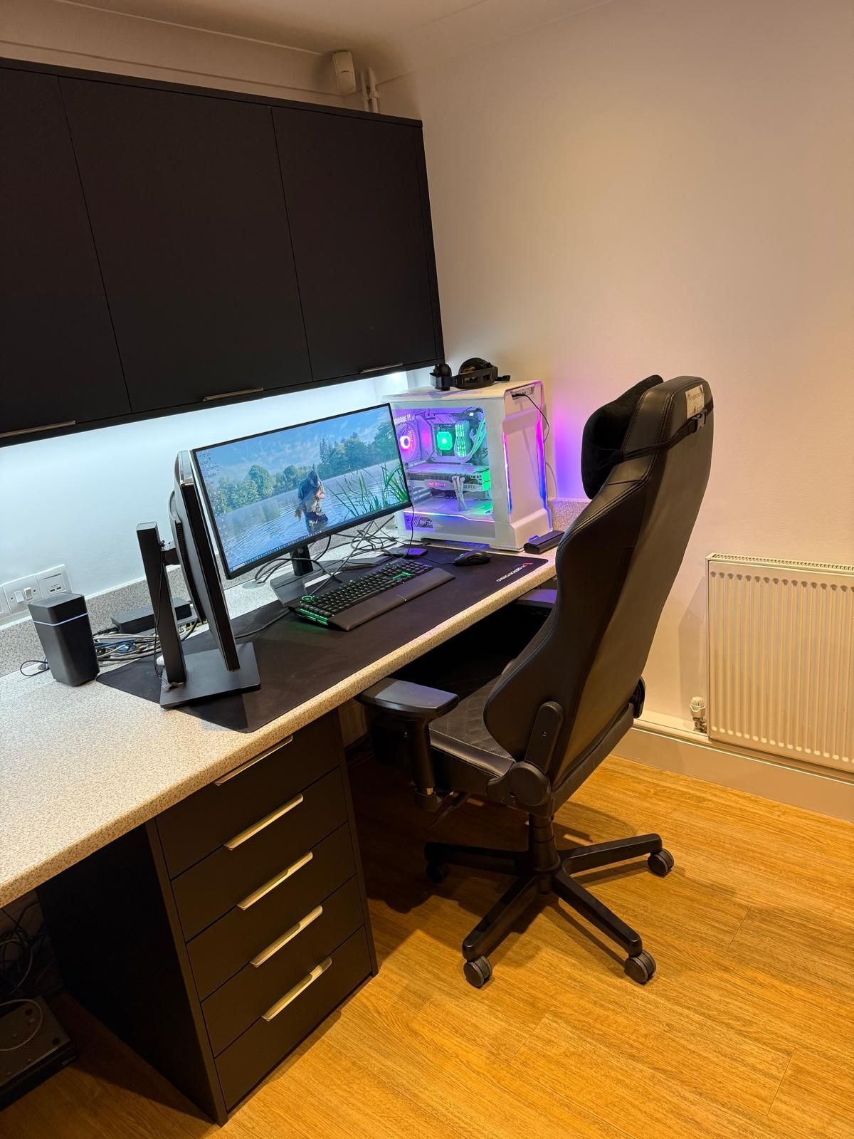 A gaming setup featuring a wide monitor, a lit PC tower, and a black gaming chair at a desk with black drawer units.