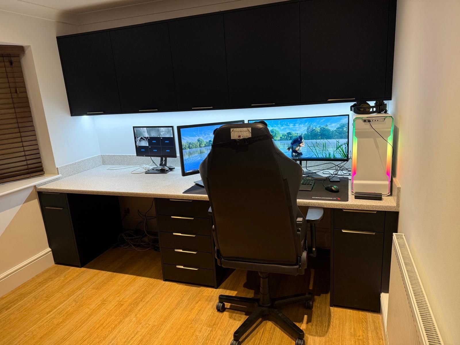 A clean home office setup featuring a large desk, ergonomic chair, dual monitors, and overhead black cabinets.