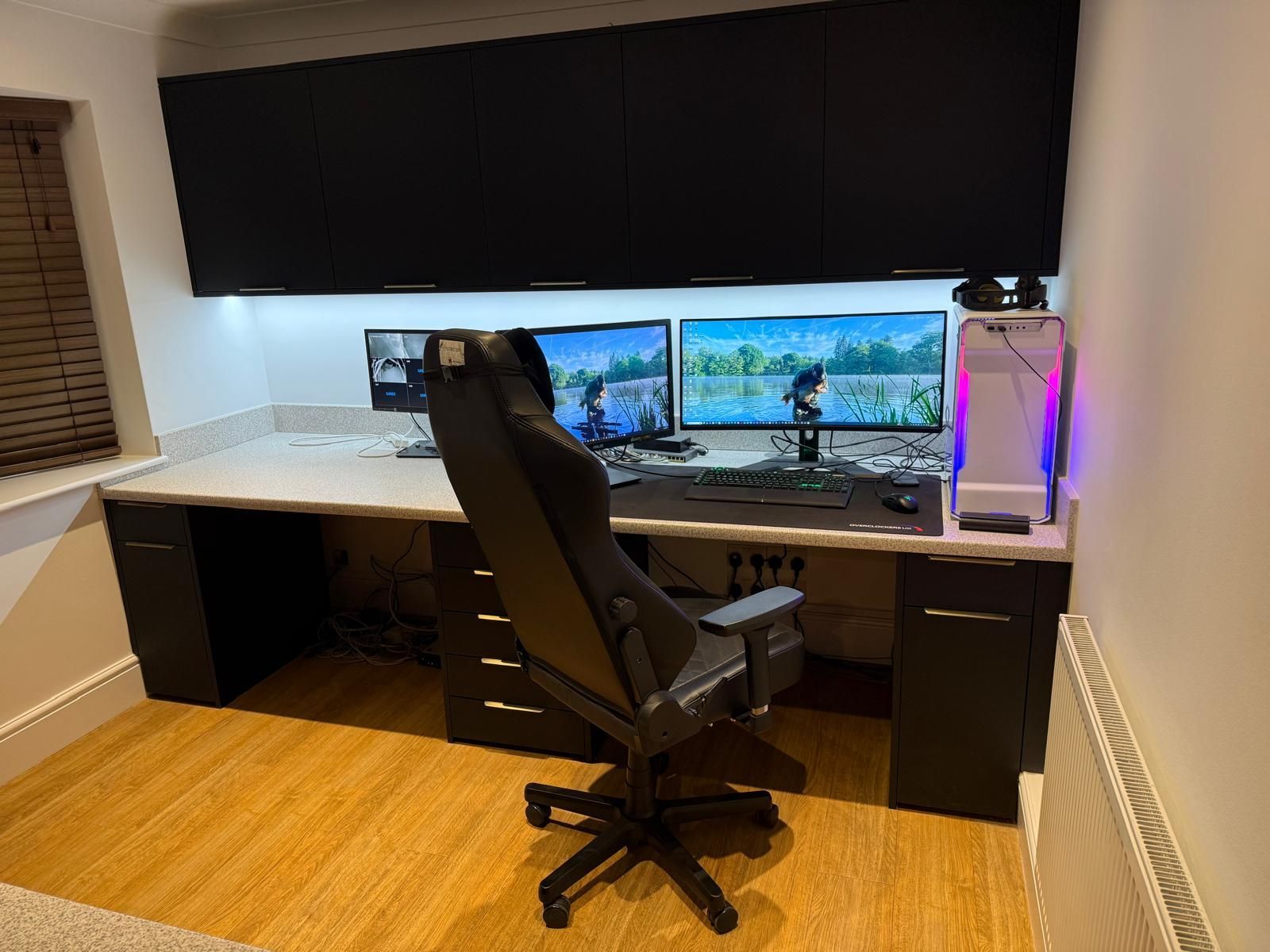 A desk setup with two monitors, a gaming chair, and black cabinets against a wall in a room with wood flooring.