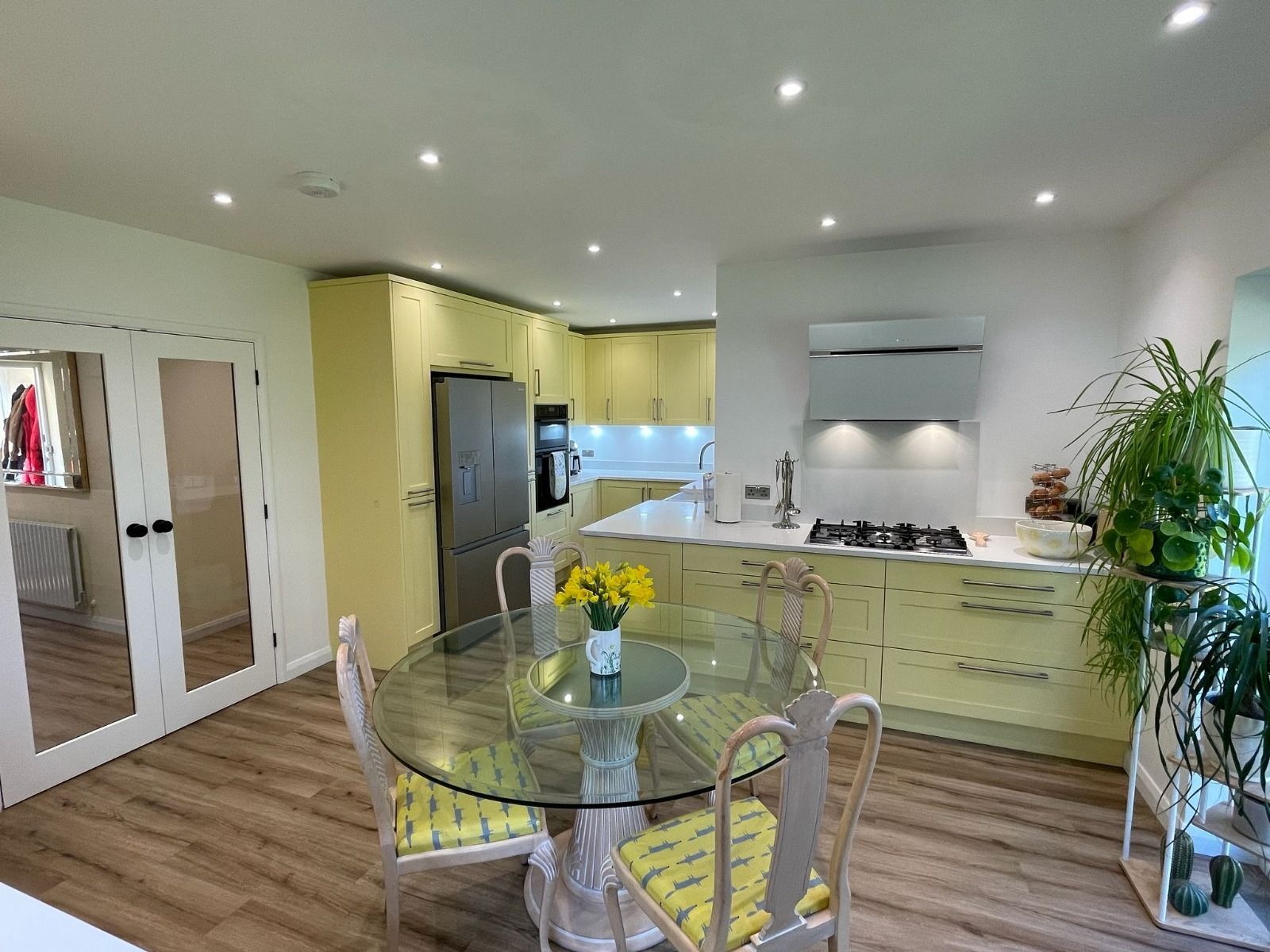A bright, modern kitchen and dining area with yellow cabinets, a glass table, four chairs, and a large potted plant.