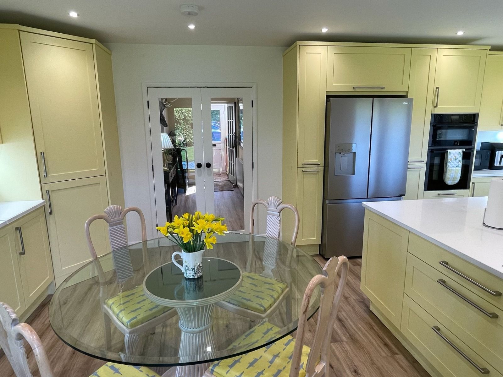 A bright kitchen featuring pale yellow cabinets, a stainless-steel refrigerator, and a circular glass dining table.