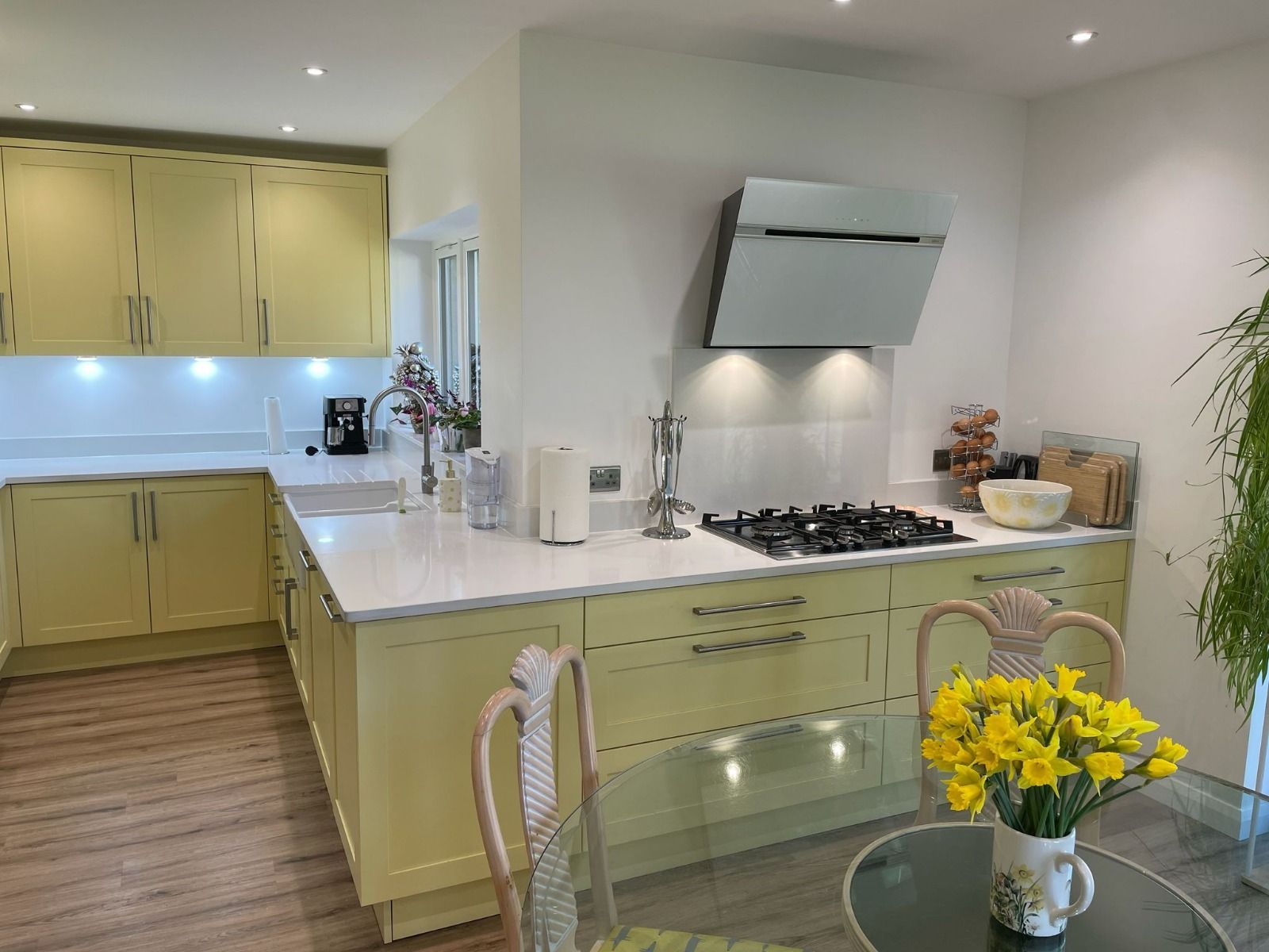 A bright kitchen features pale yellow cabinets, white countertops, a gas cooktop with a vent hood, and a small dining area.