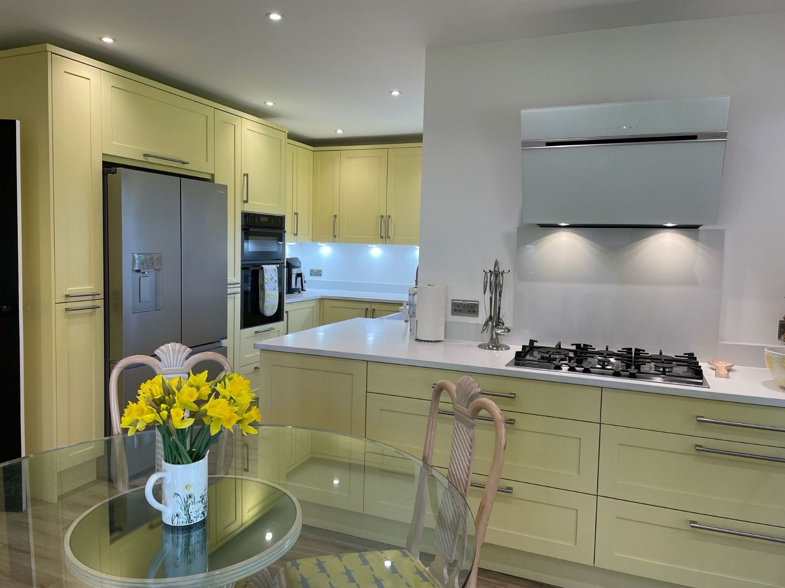 A modern kitchen with light yellow cabinets, stainless steel appliances, a glass table with daffodils, and white counters.
