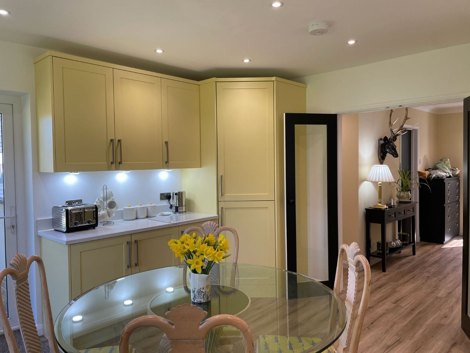 A sunny yellow kitchen with a glass round table, chairs, a toaster, and a doorway leading to a hallway with a stag head.