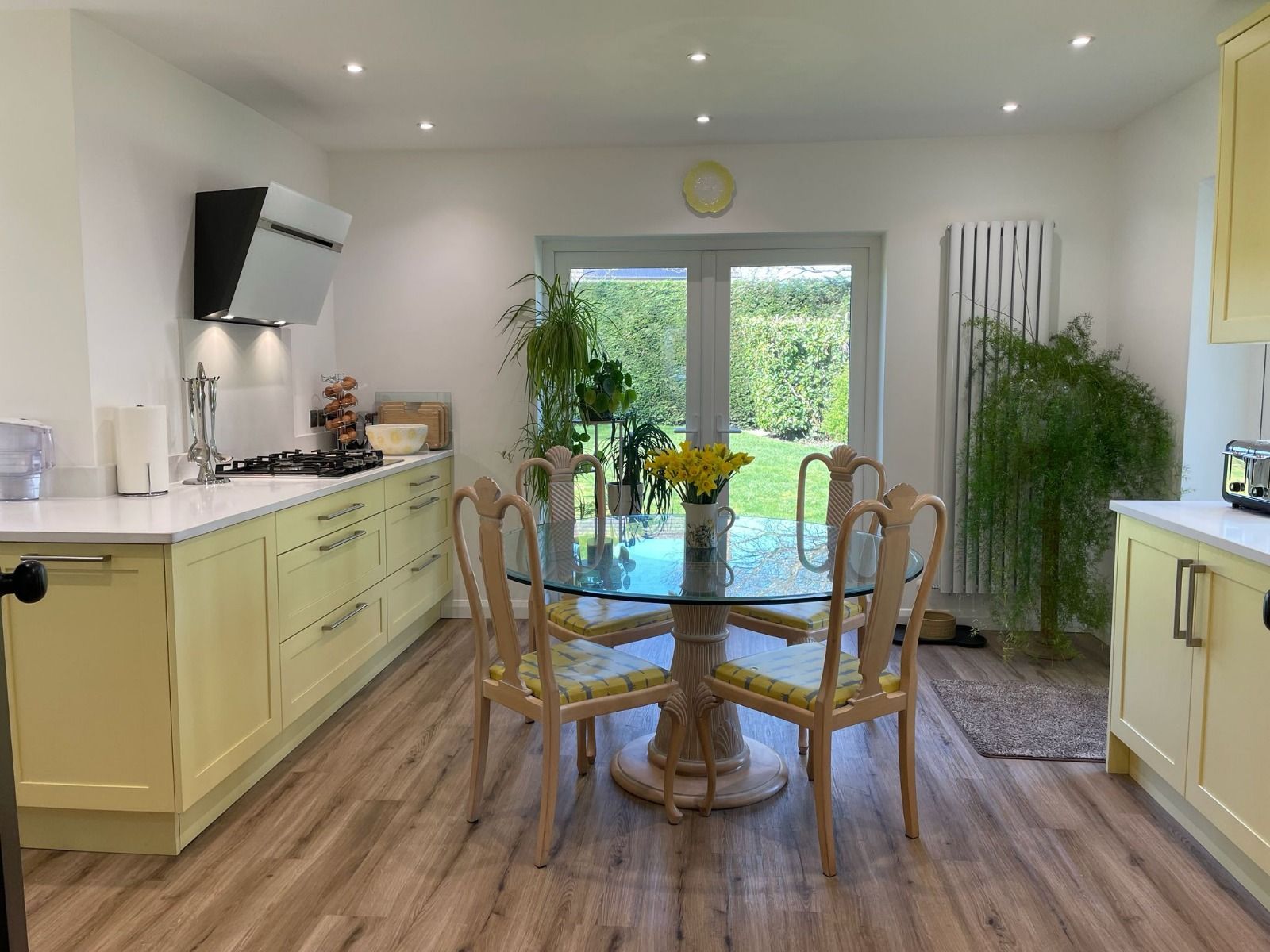 A bright kitchen with light yellow cabinets, a round glass dining table, wood-look flooring, and a view of a garden.