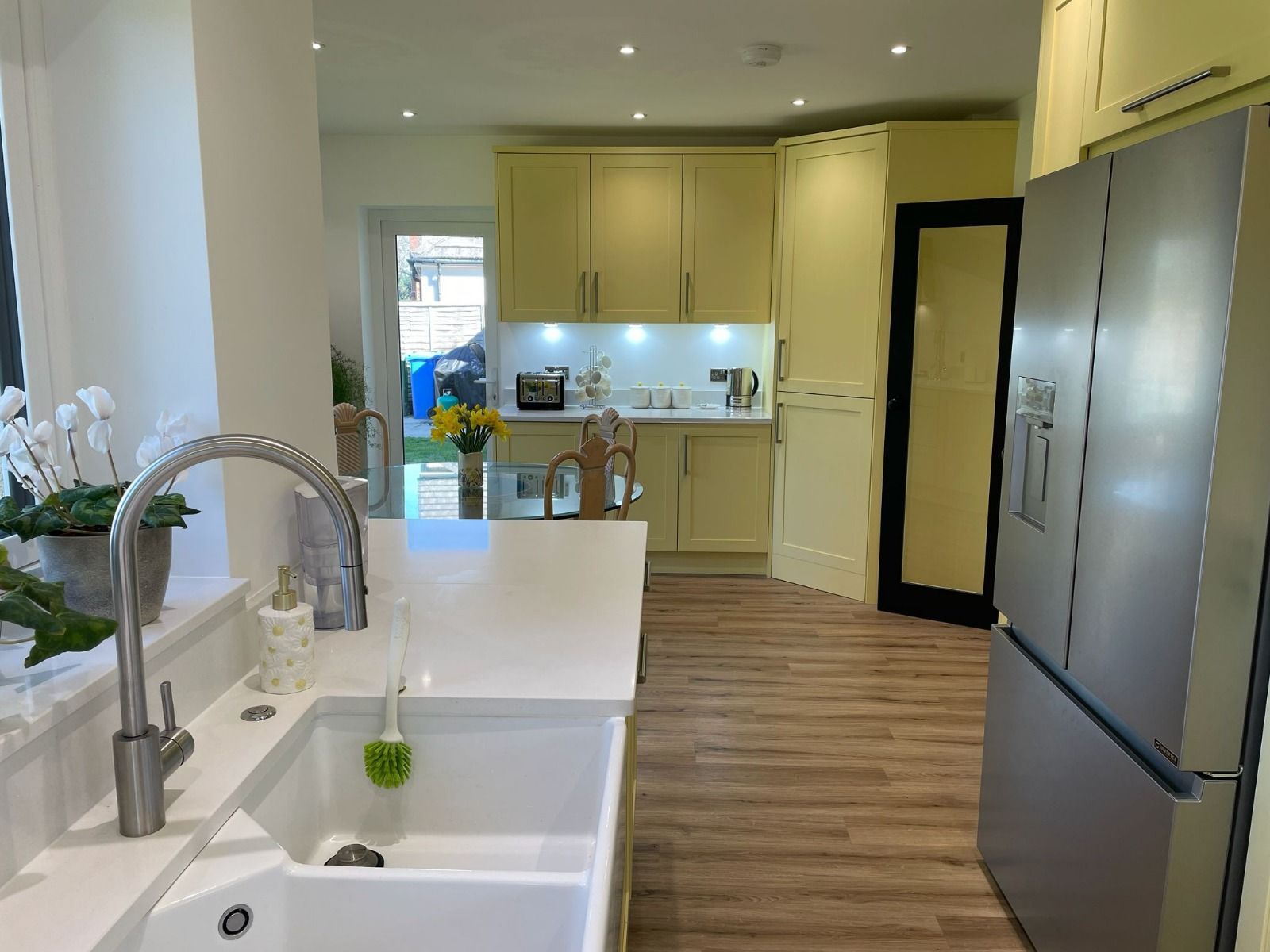 A modern kitchen with light yellow cabinets, white countertops, a large farmhouse sink, and a stainless steel refrigerator.