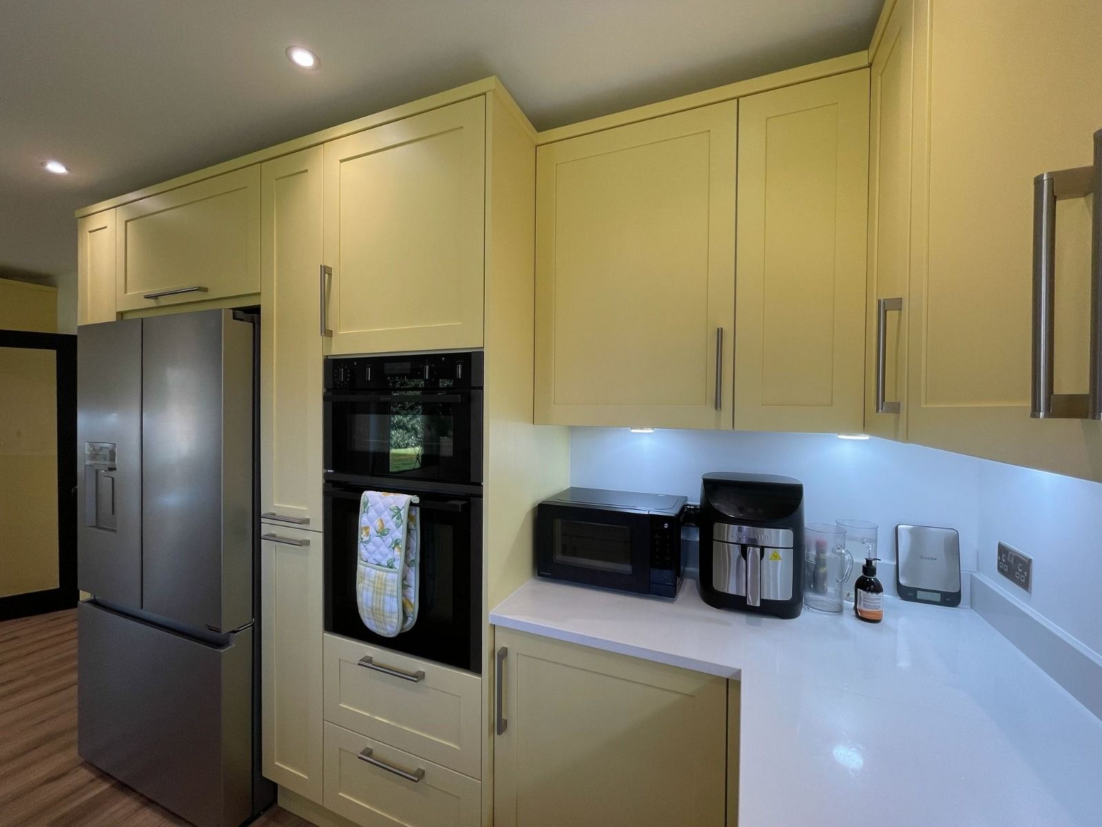 A kitchen with pale yellow cabinets, stainless steel appliances, and white countertops with under-cabinet lighting.