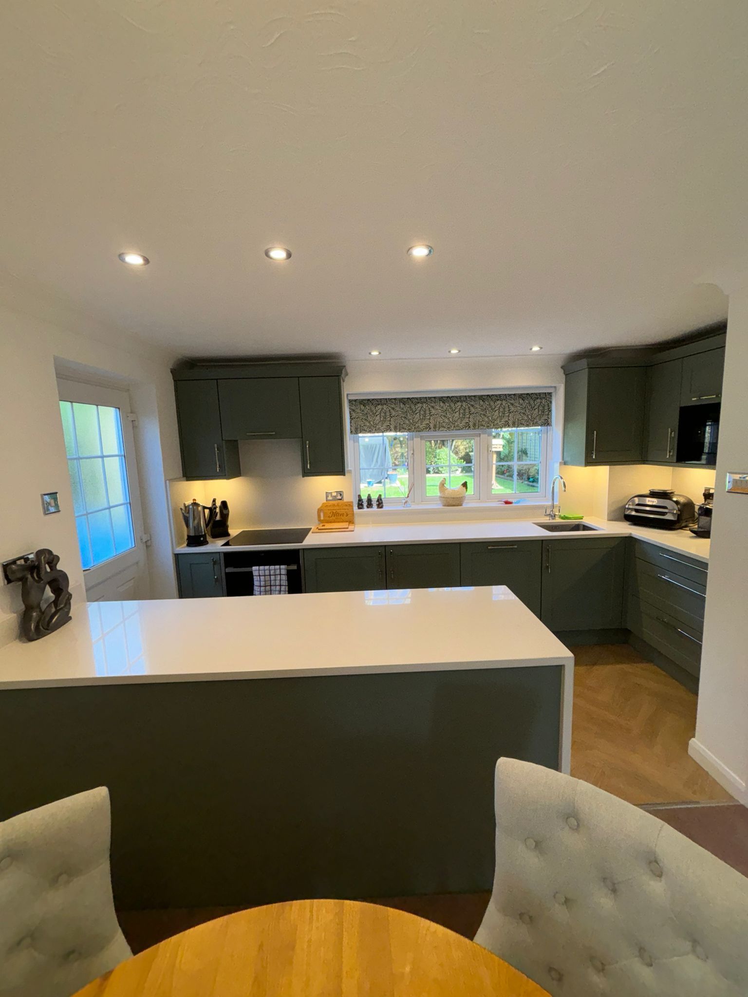 A modern kitchen with dark green cabinets, white countertops, a central island, and a window overlooking a garden.
