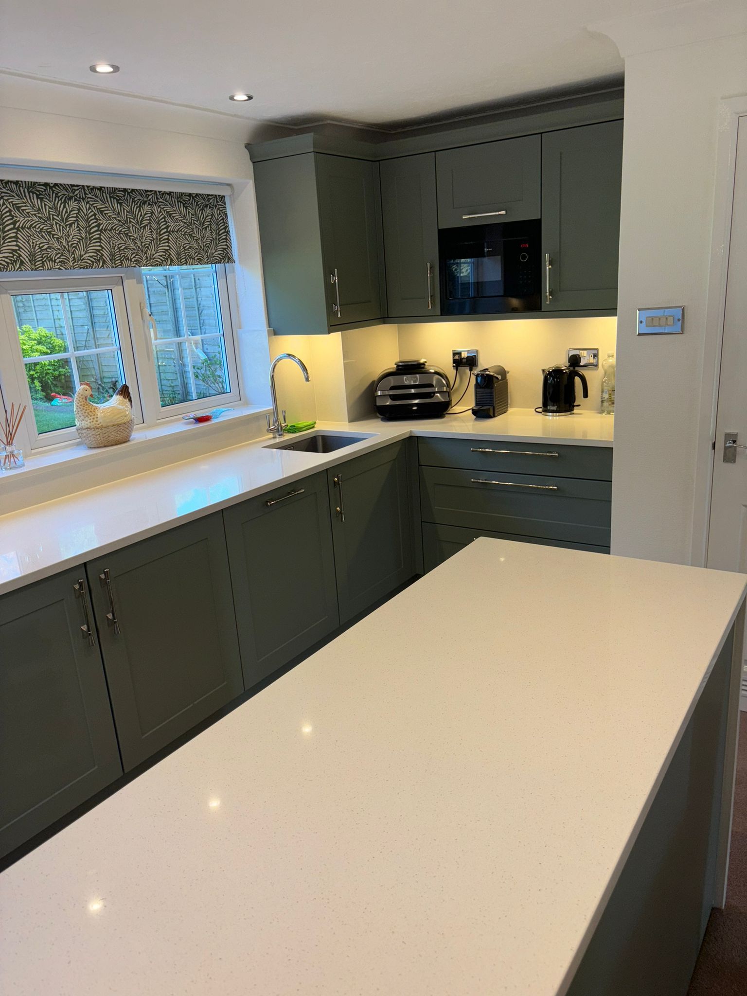 A kitchen with sage-green cabinets, white quartz countertops, an island, and a window with a patterned valance.