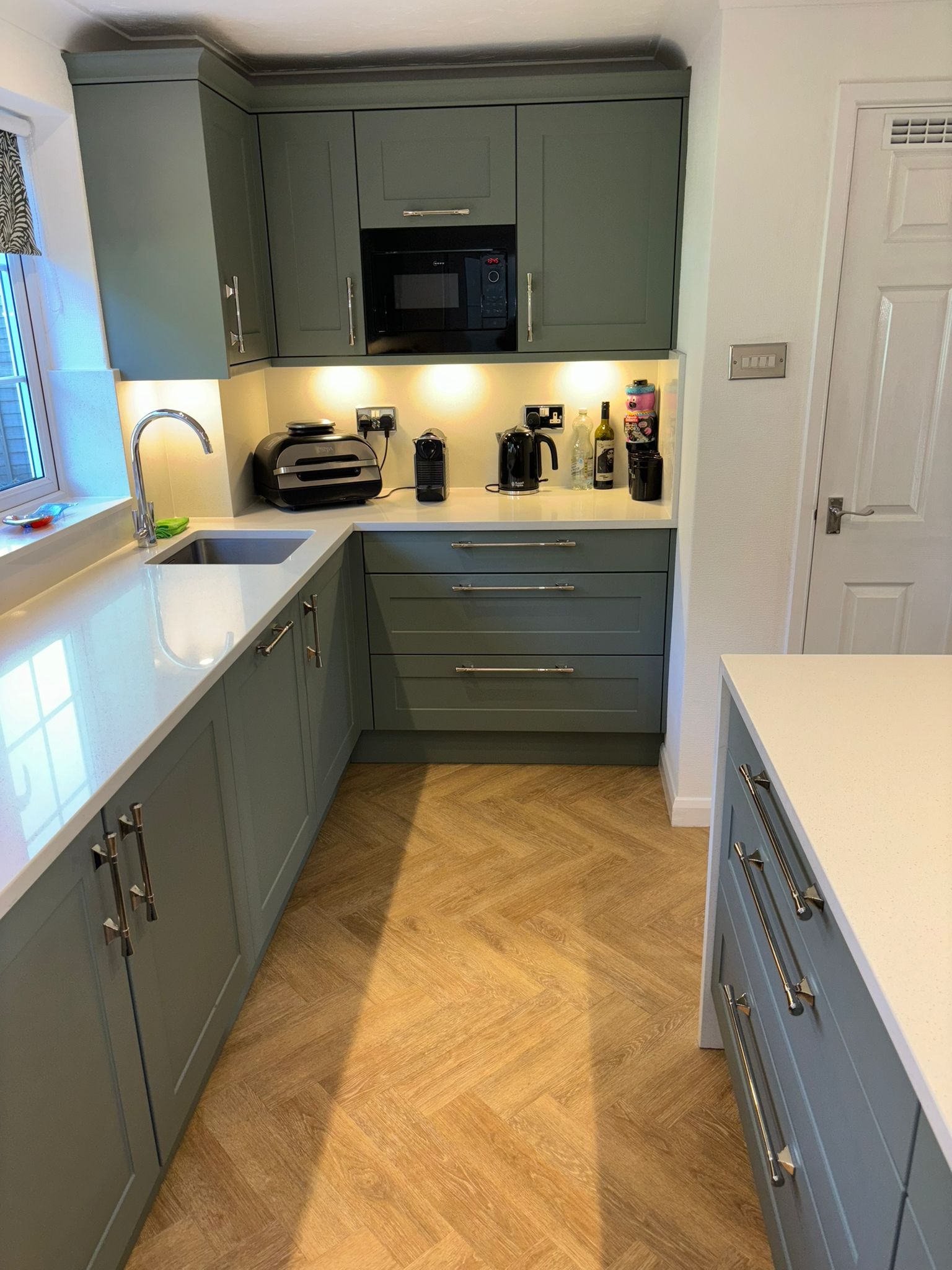 A modern galley-style kitchen featuring sage green cabinets, white countertops, and light brown herringbone flooring.