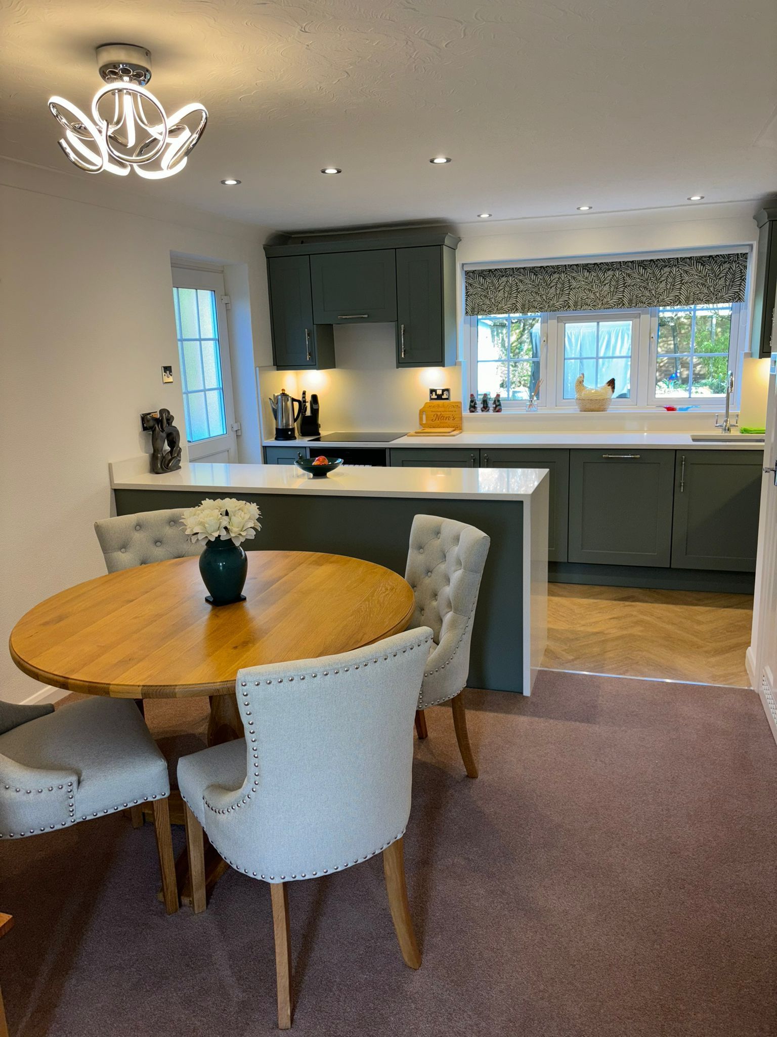 Dining room with a round wooden table, four tufted upholstered chairs, and an open kitchen with dark grey cabinets.