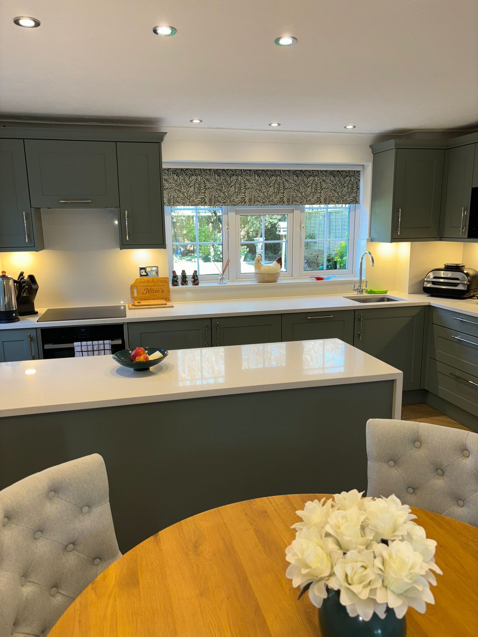 A modern kitchen with sage green cabinets, white countertops, a central island, and a dining table with white flowers.