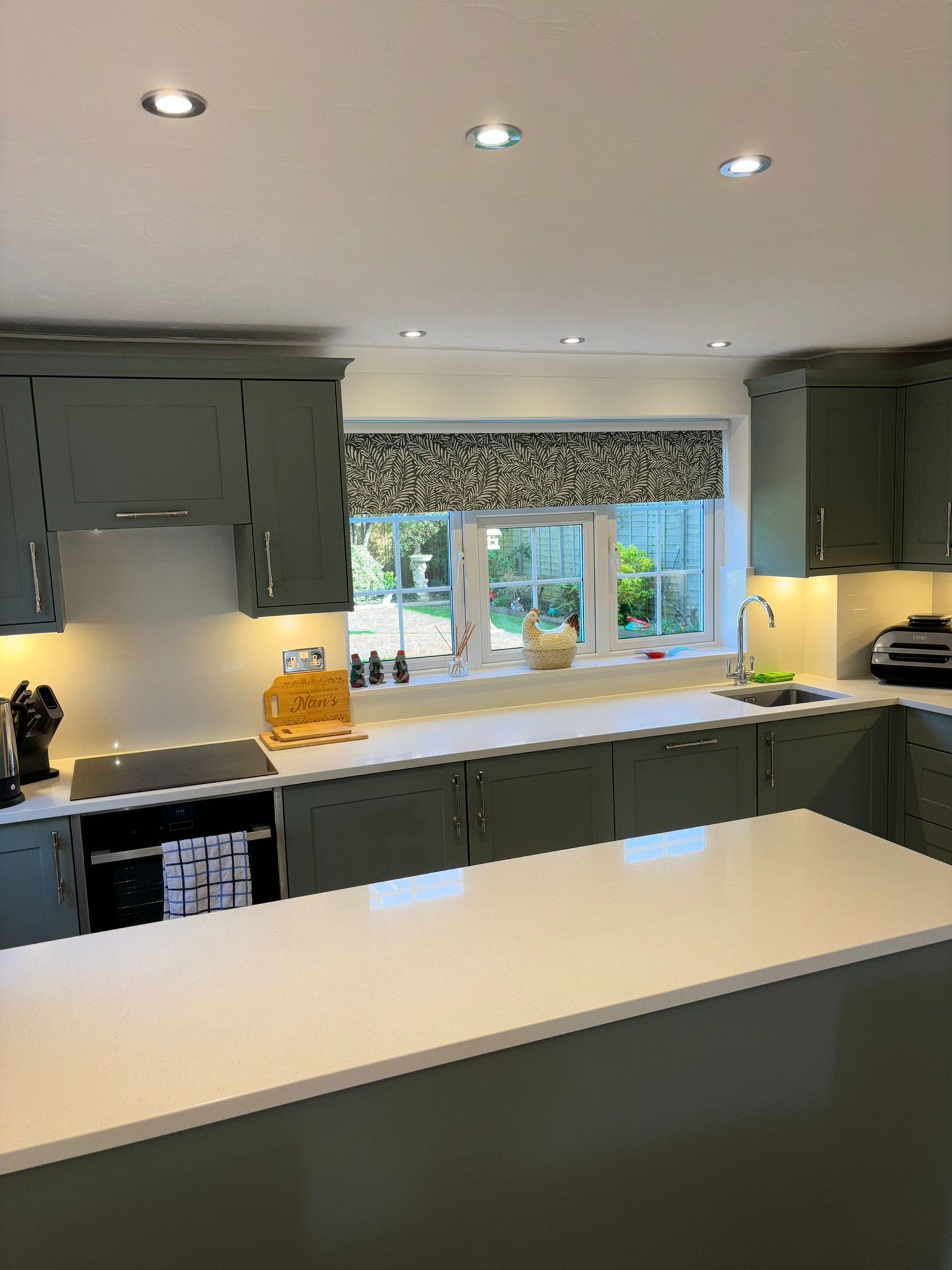 Modern kitchen with sage green cabinets, white countertops, a central island, and a patterned Roman blind on the window.