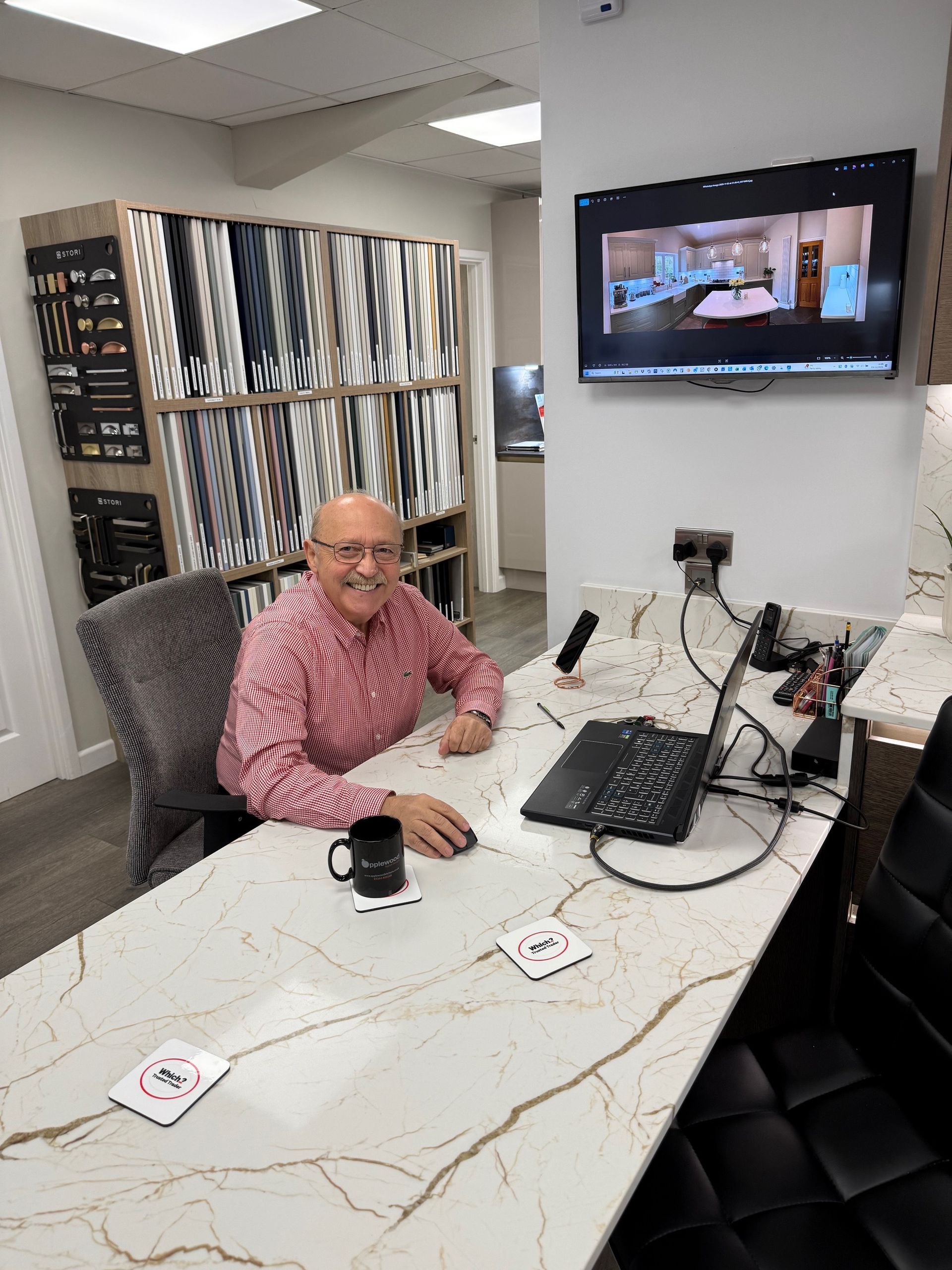A person sits at a marble desk in an office with fabric samples on shelves and a large screen displaying a room interior.