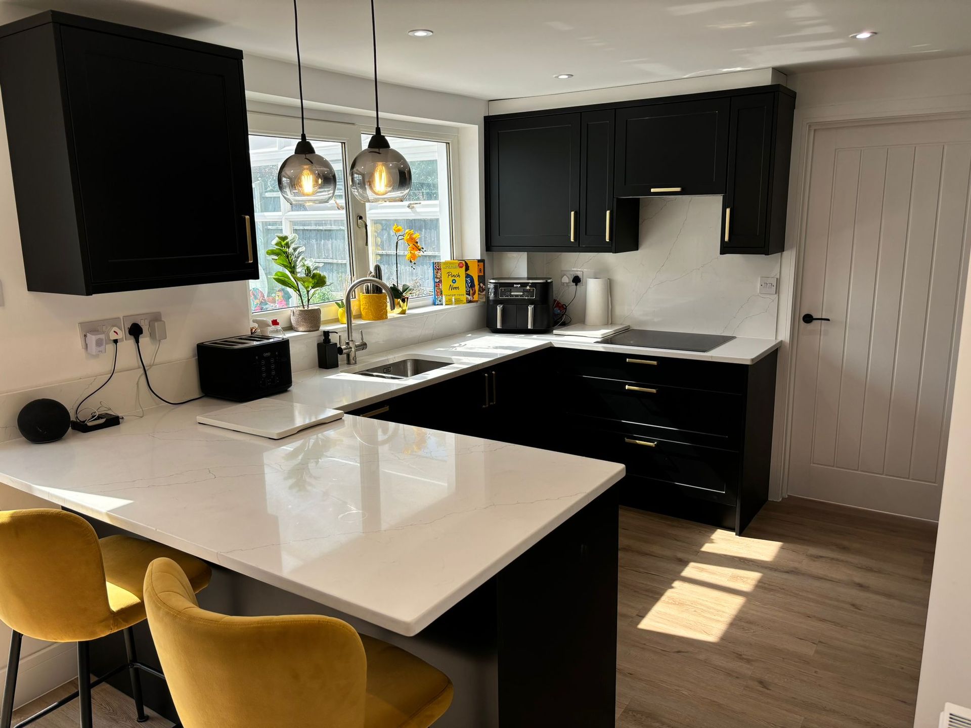 A modern kitchen with black cabinets, white countertops, two yellow bar stools, and pendant lighting over a peninsula.
