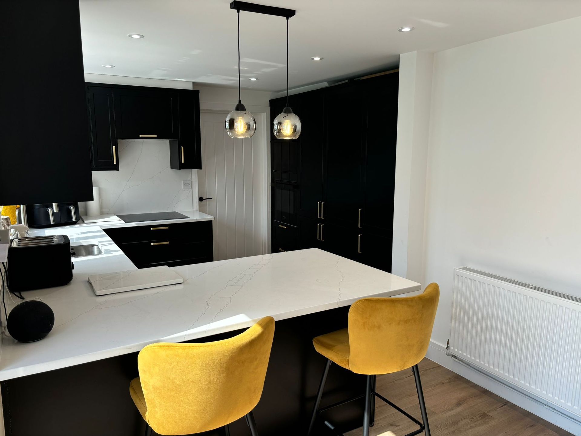 A modern kitchen featuring black cabinetry, a white countertop island, two yellow velvet bar stools, and pendant lighting.