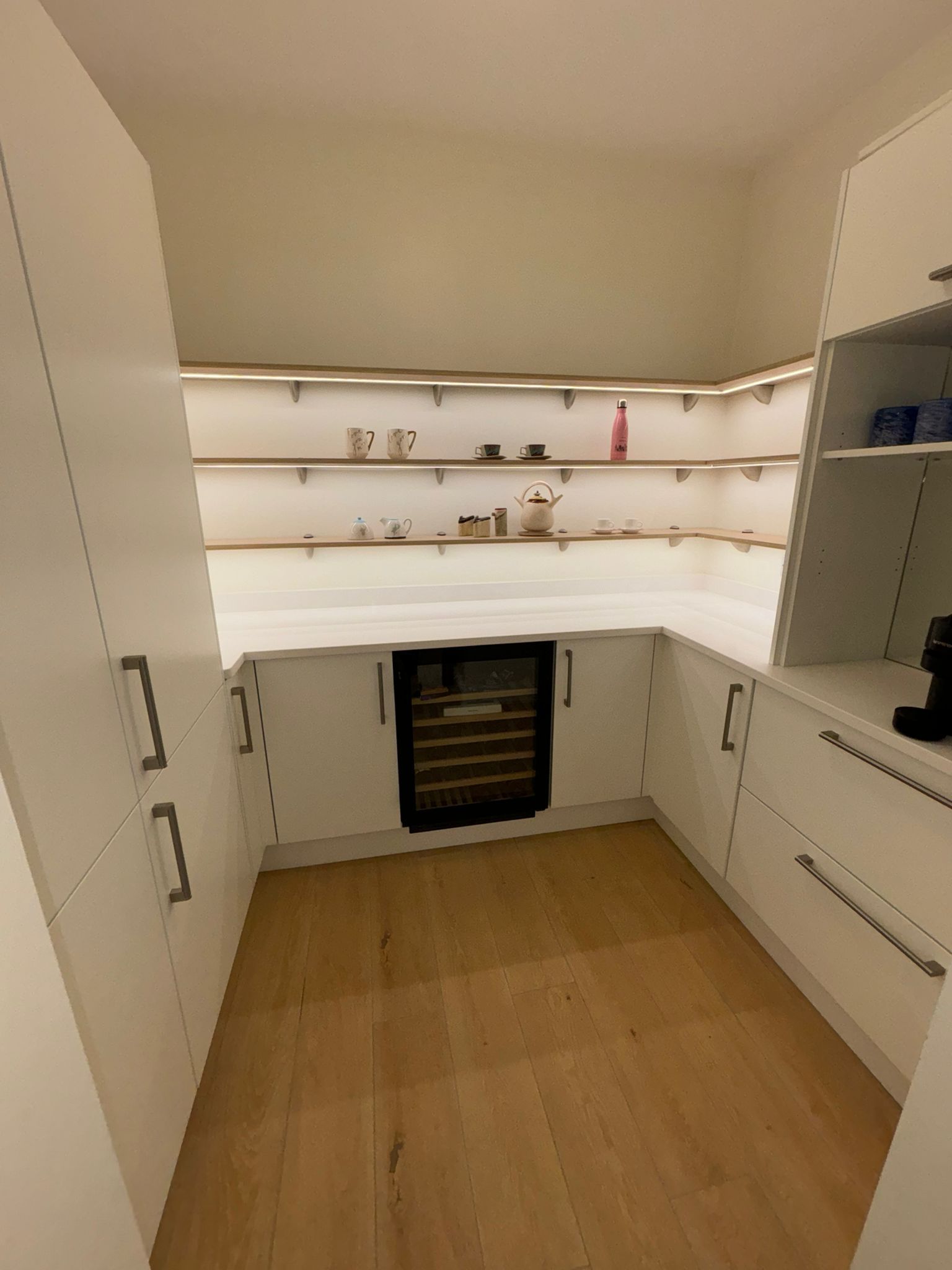 A bright, modern white pantry featuring open shelving with under-cabinet lighting, a wine fridge, and wood flooring.