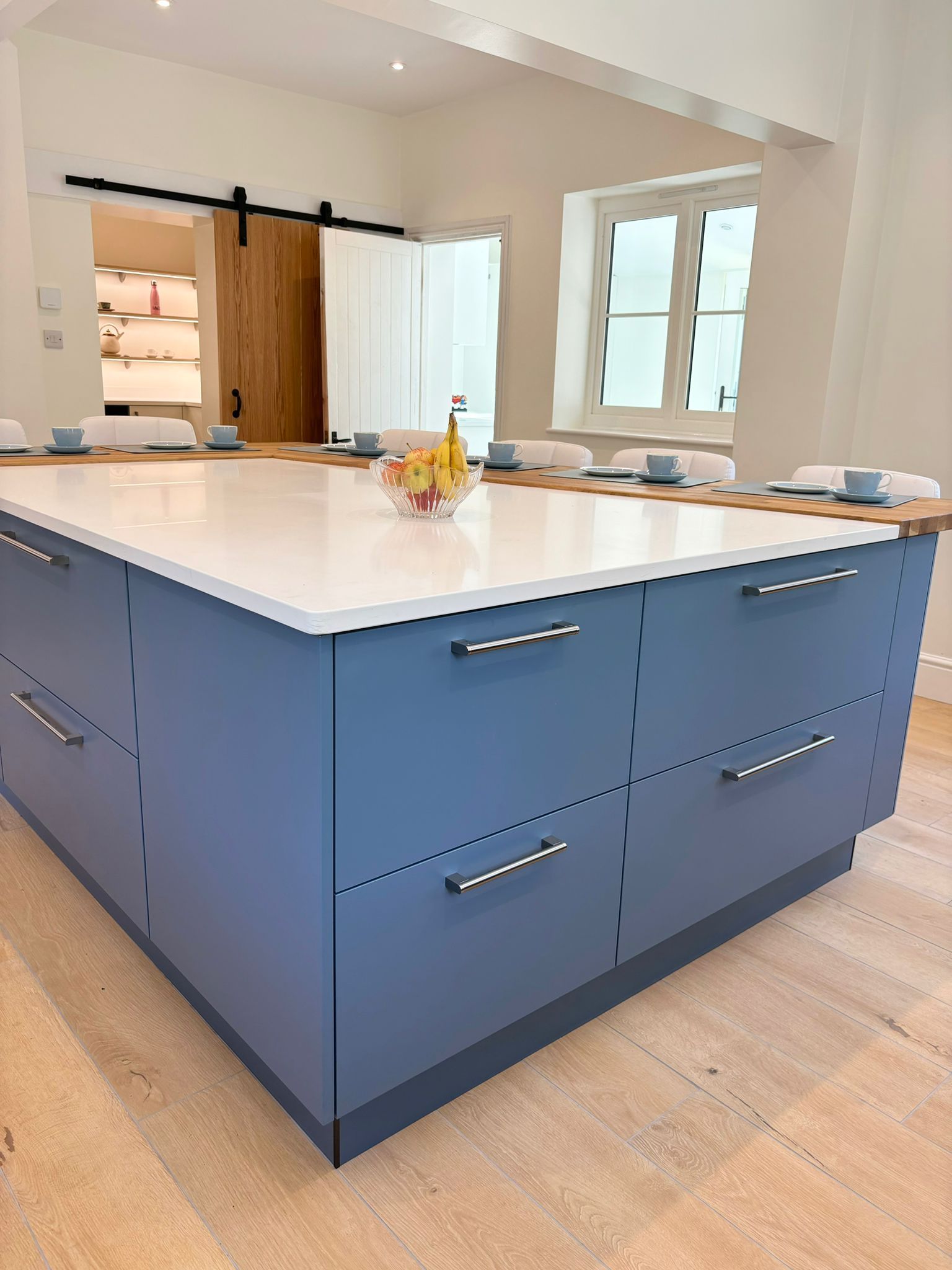 A large, central kitchen island with blue cabinets, a white countertop, and metal pulls sits on a light wooden floor.