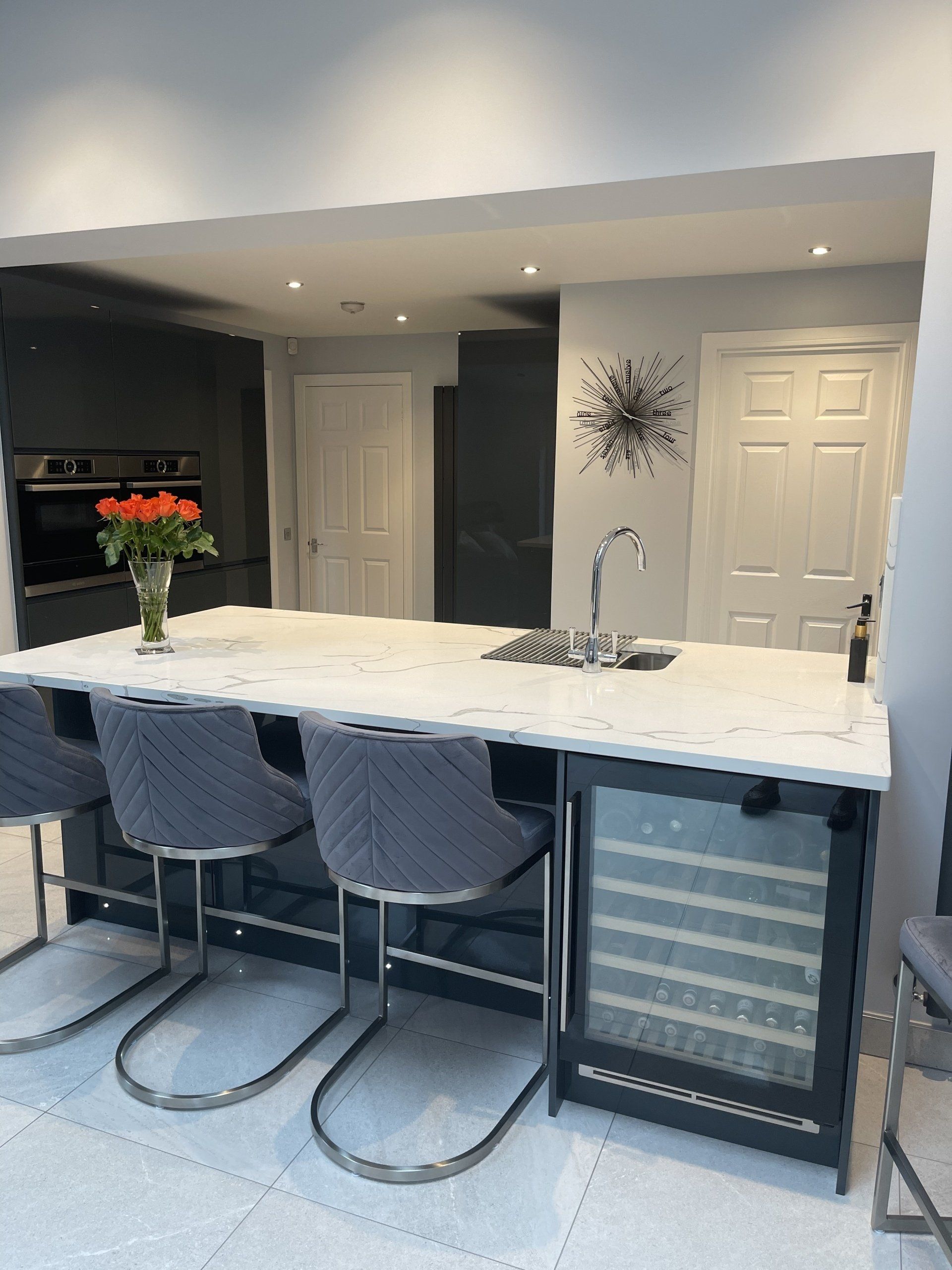 A kitchen island with a white marble countertop, a built-in wine fridge, and grey velvet bar stools in a modern kitchen.