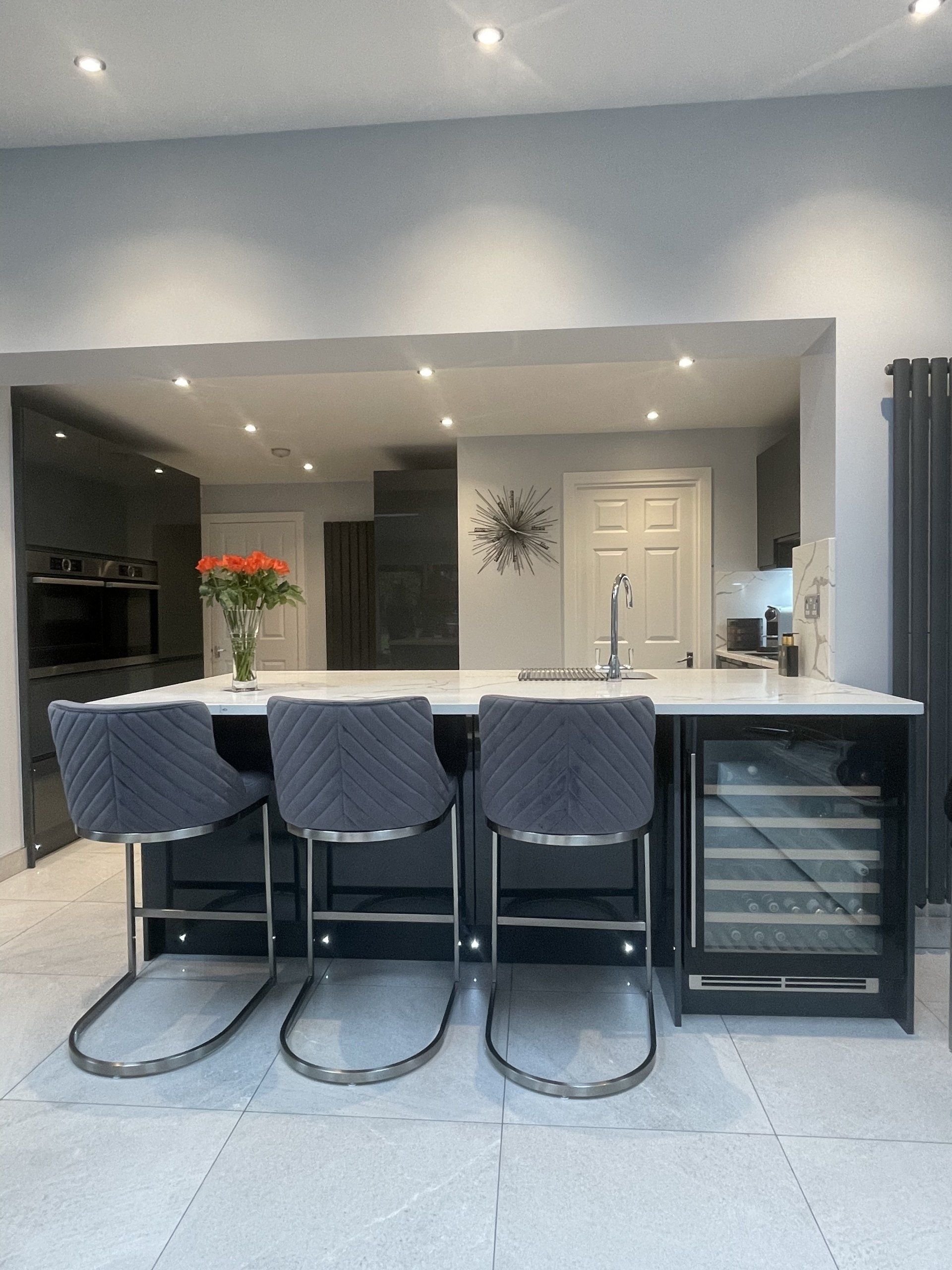 A modern kitchen island with three grey quilted bar stools and a built-in wine cooler, featuring a vase of orange flowers.