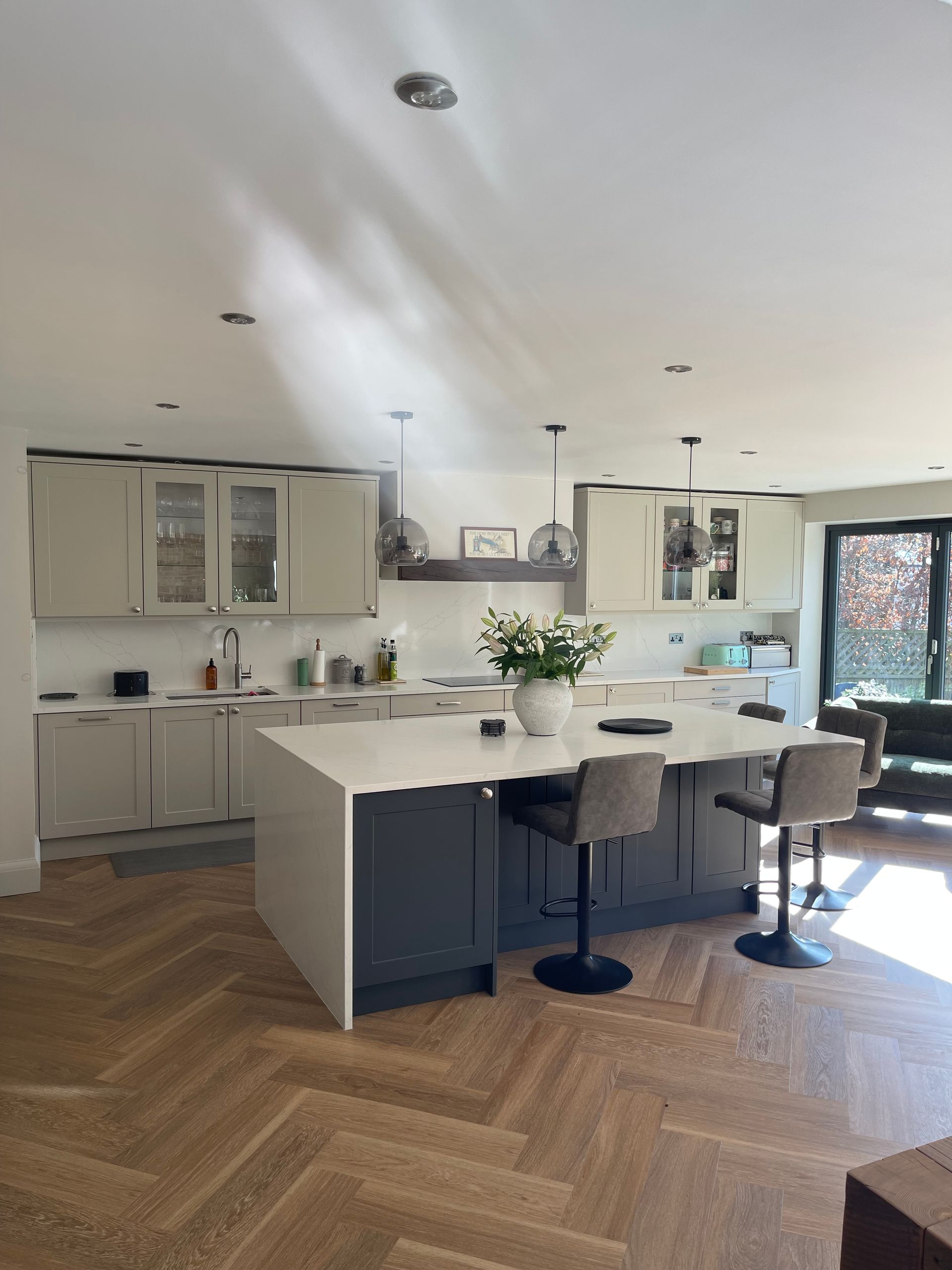 A modern kitchen featuring a dark grey central island, light grey cabinets, white countertops, and herringbone flooring.