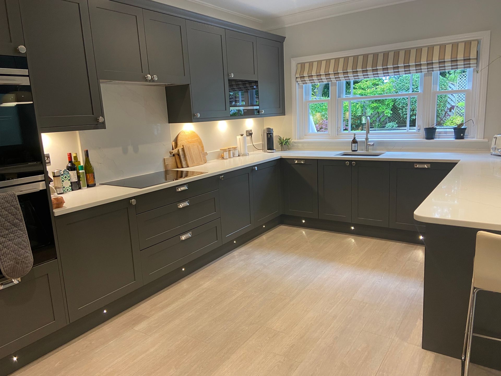 A modern L-shaped kitchen featuring charcoal cabinets, white countertops, stainless steel appliances, and wood flooring.