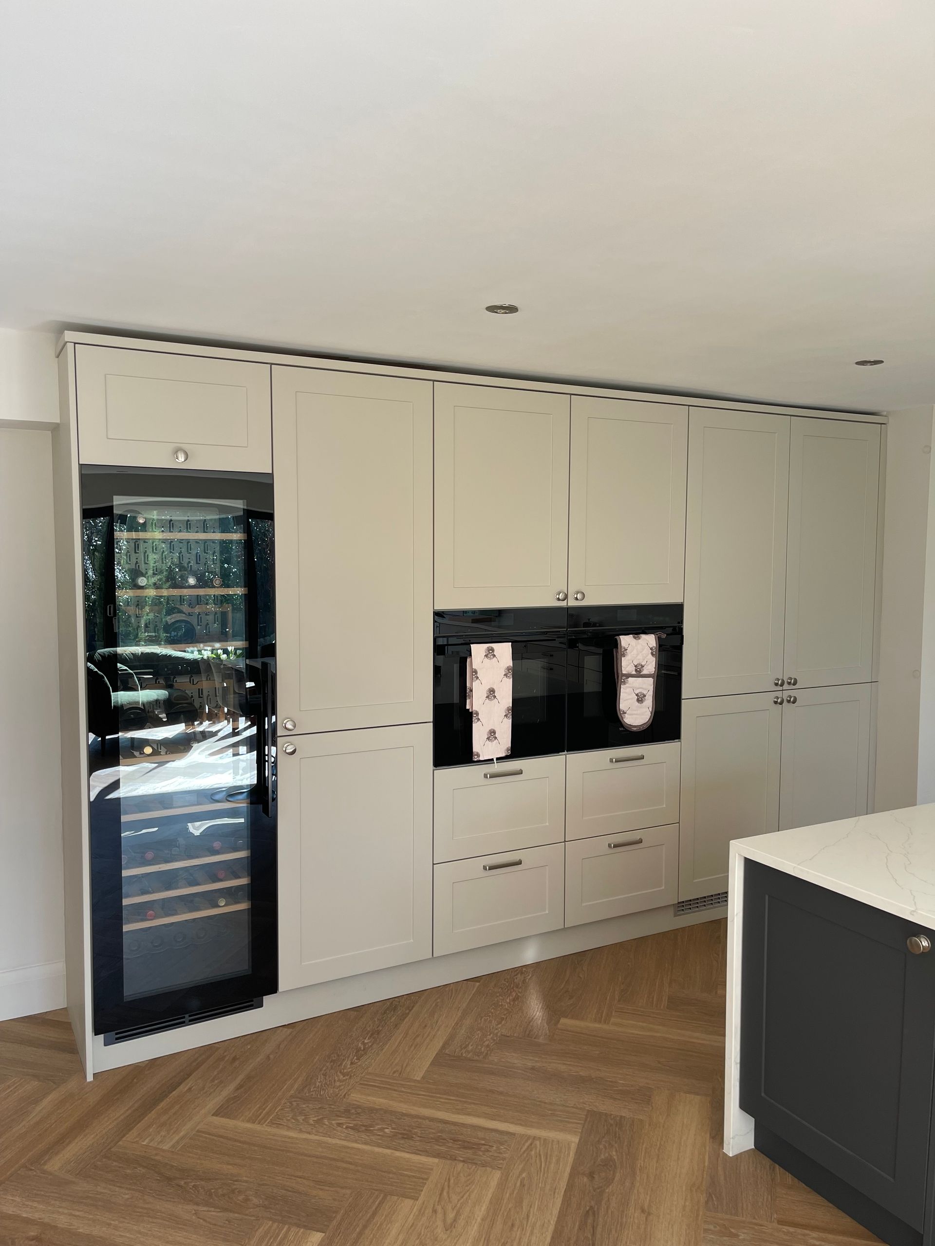 A modern wall of off-white kitchen cabinetry featuring a built-in wine cooler, two ovens, and herringbone wood flooring.