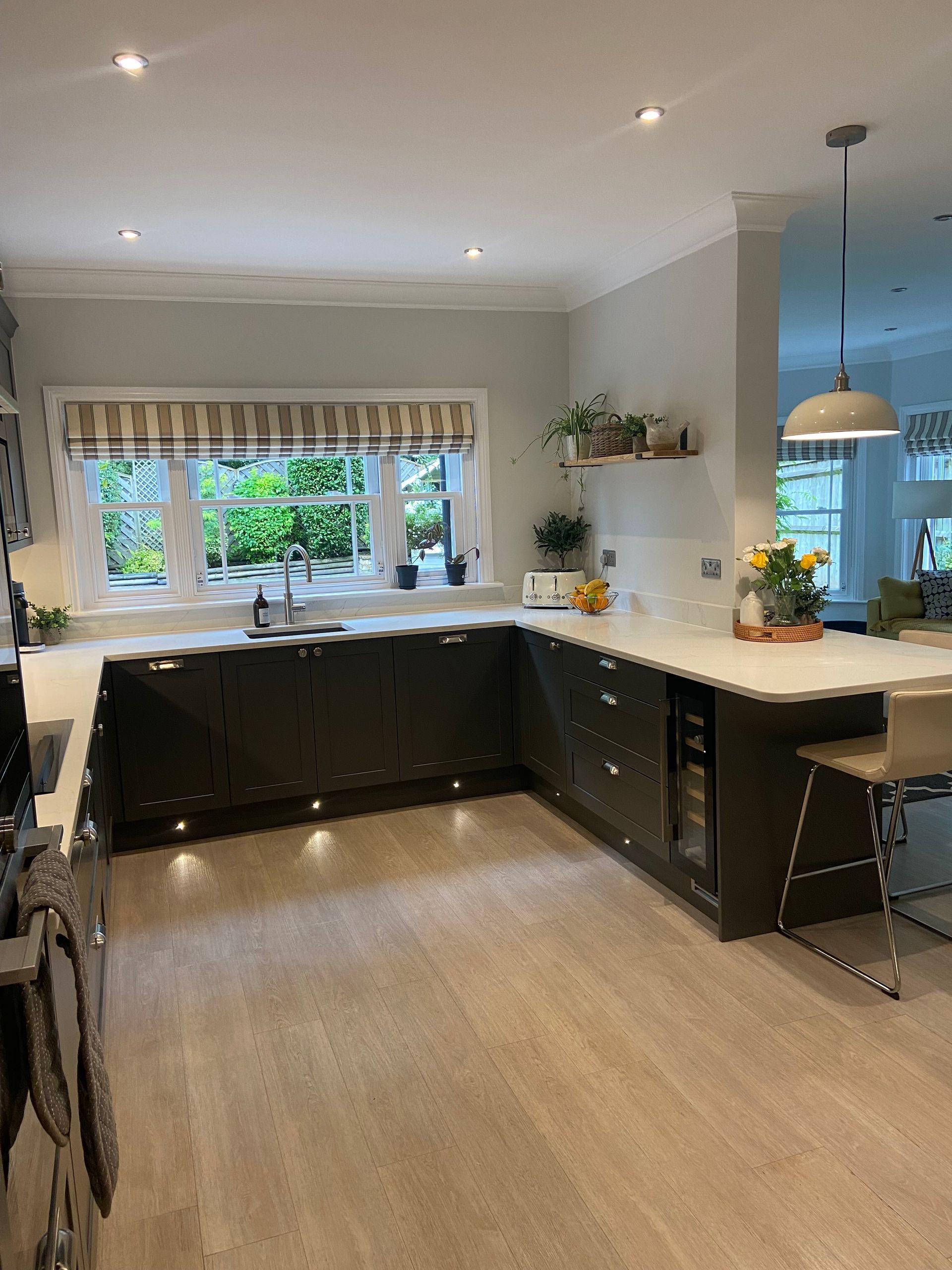 A modern kitchen with dark green cabinetry, white countertops, light wood floors, and a large window above the sink.