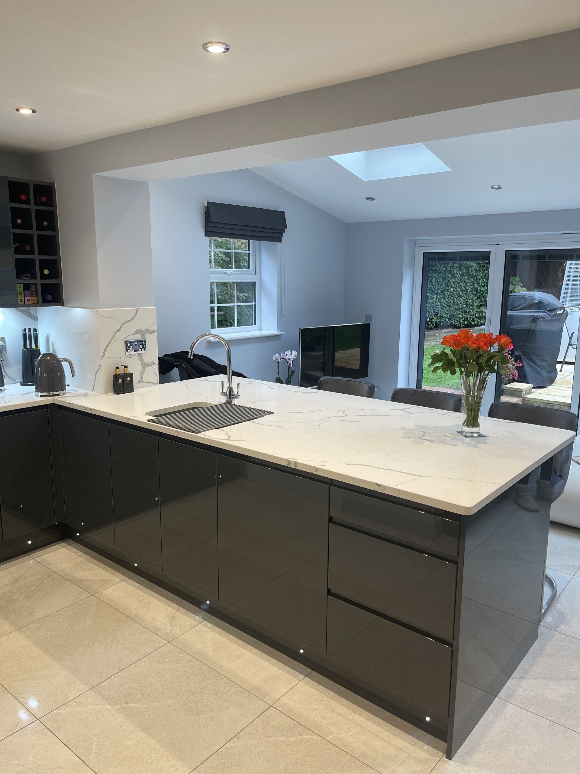 Modern kitchen with a dark grey island, white countertop, sink, and tile flooring, opening into a sunlit living space.