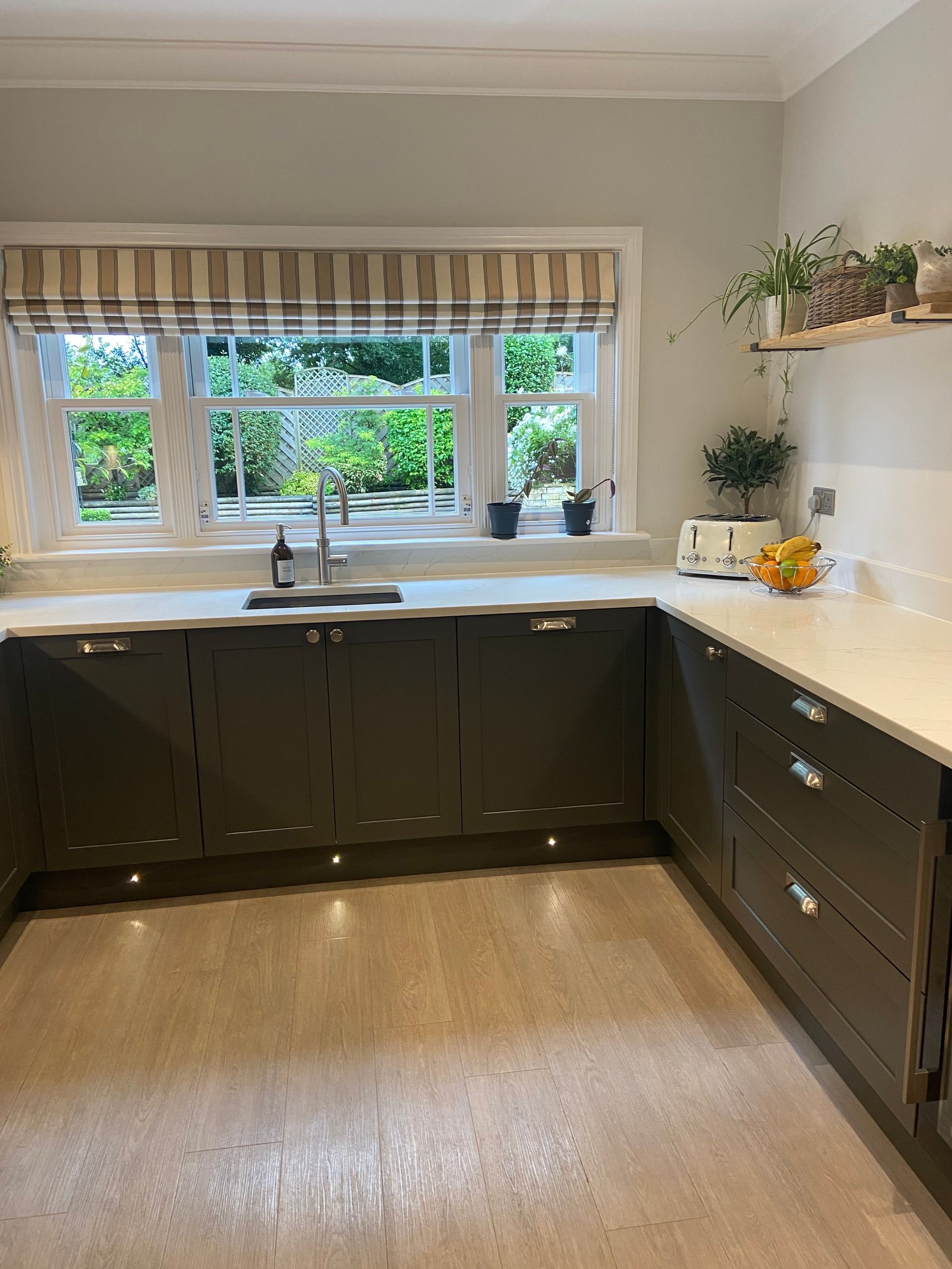 Modern L-shaped kitchen featuring charcoal cabinets, white countertops, a window with striped Roman blinds, and shelving.