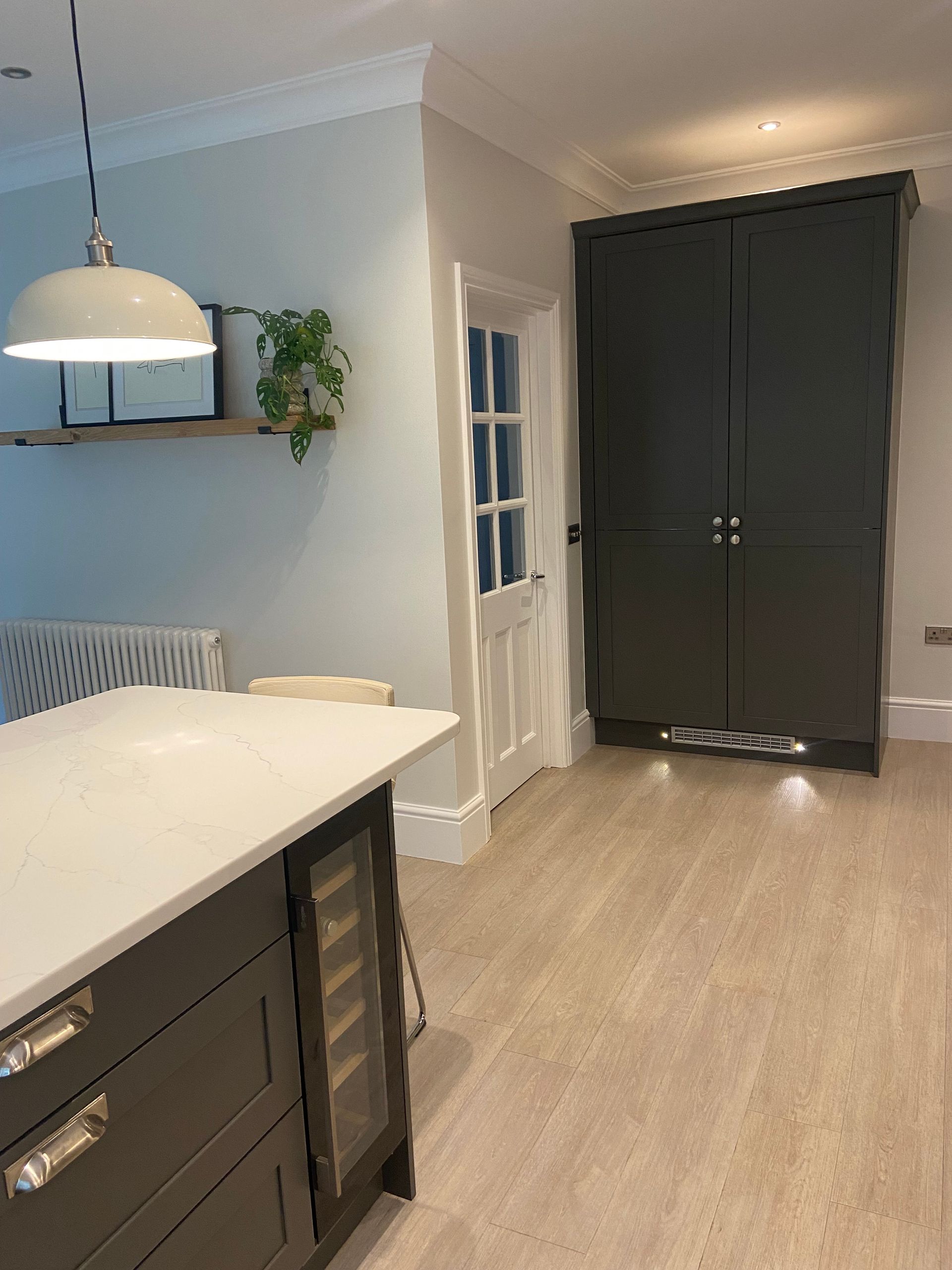 A kitchen featuring a dark gray cabinet, white countertop island with a wine cooler, a white door, and hanging pendant light.