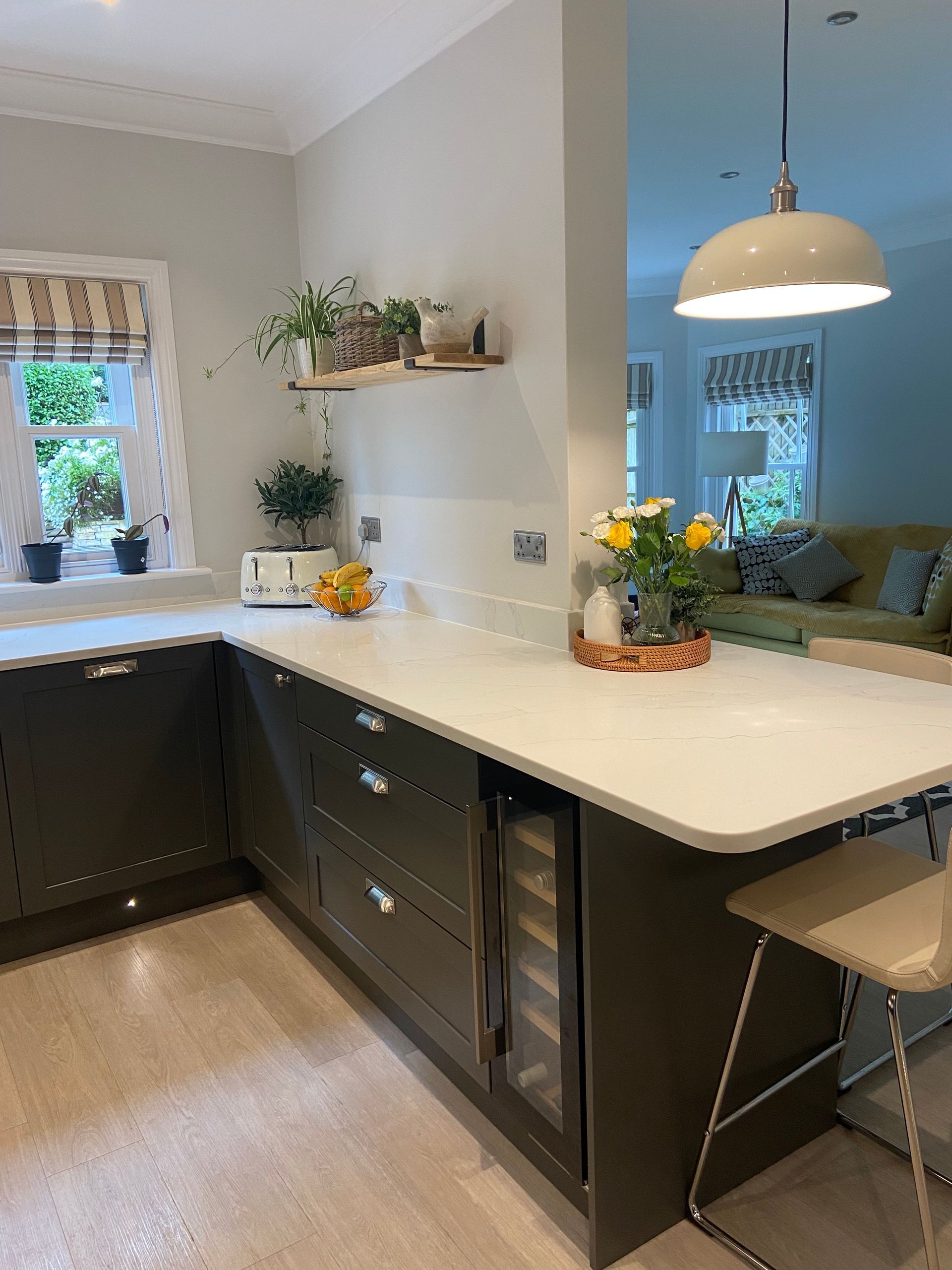 A modern kitchen with dark grey cabinets, white countertops, a wooden shelf with plants, and a view into a living room.