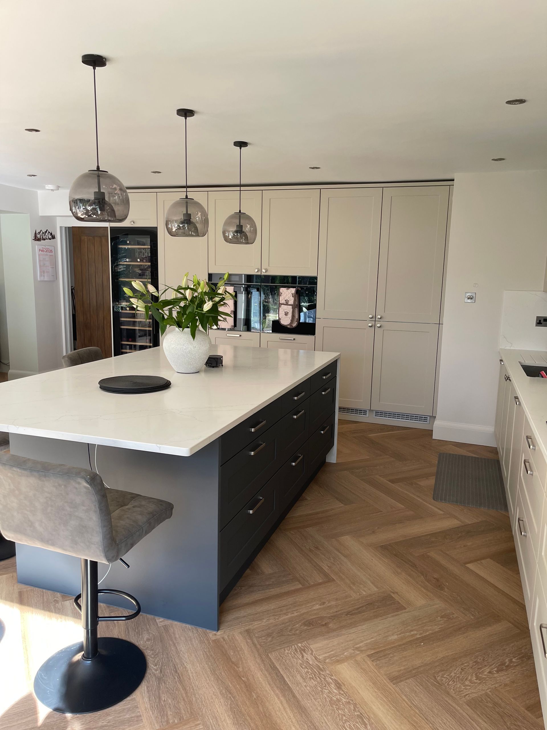 A modern kitchen with a large dark blue island, white countertops, three pendant lights, and herringbone wood flooring.