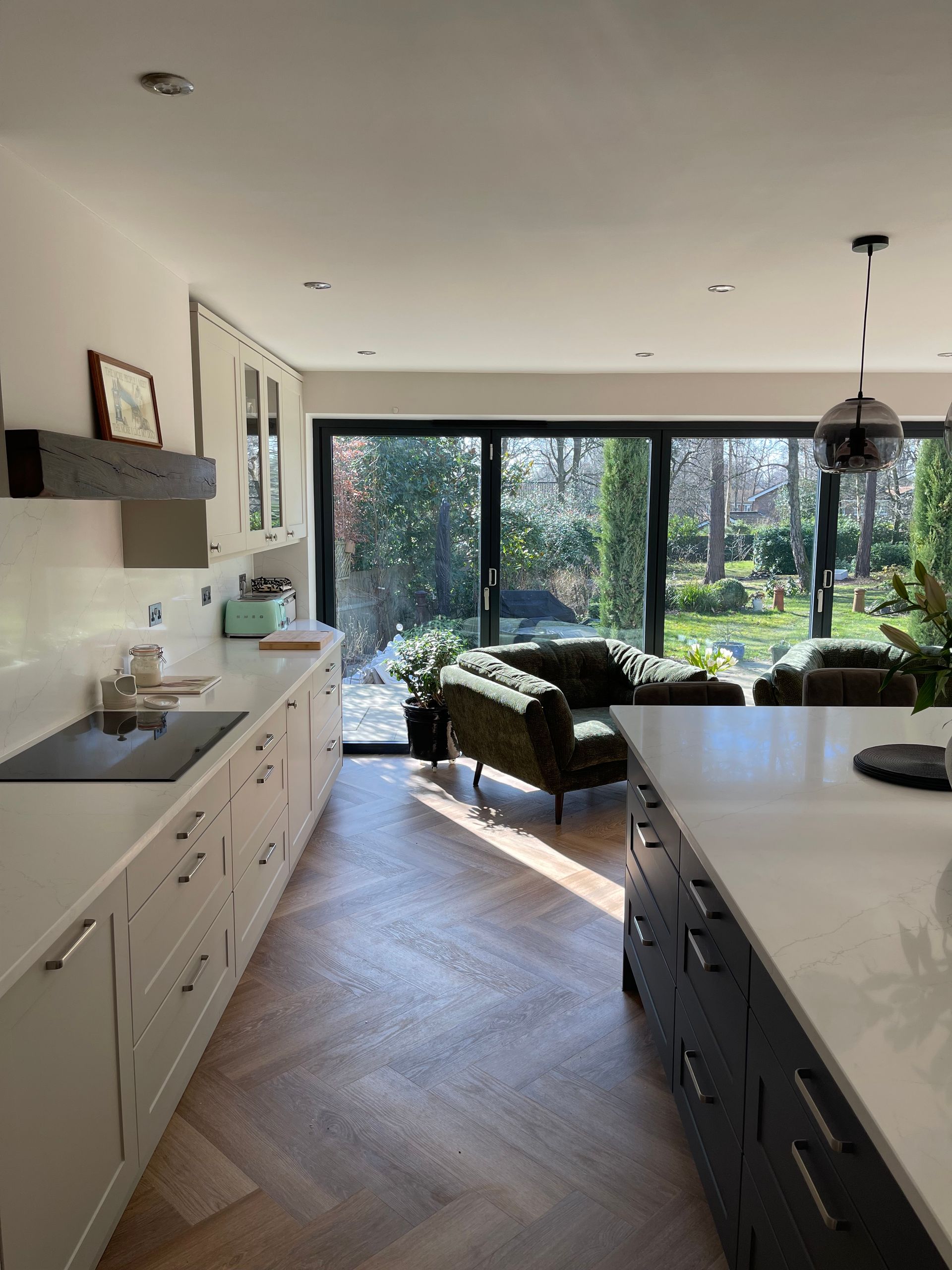A bright kitchen with white cabinets, a dark island, herringbone wood floors, and large glass doors overlooking a yard.