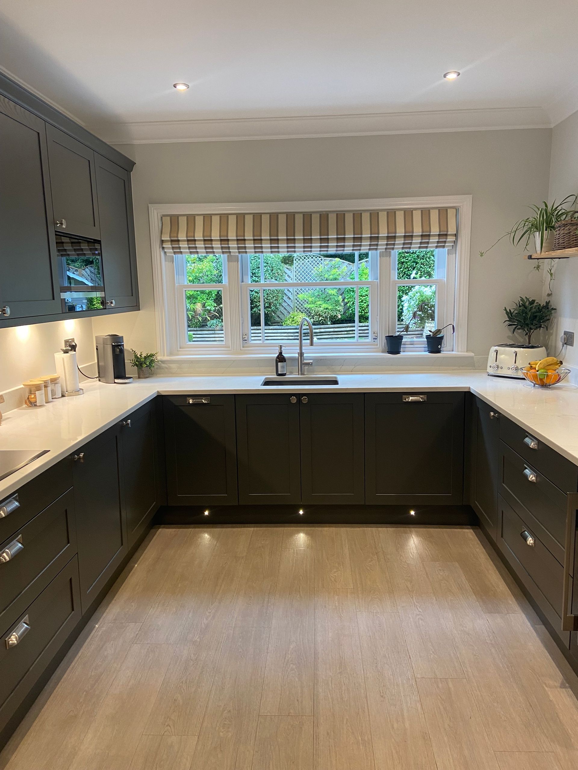 A U-shaped kitchen with dark grey cabinets, white countertops, a large window over the sink, and light-toned flooring.