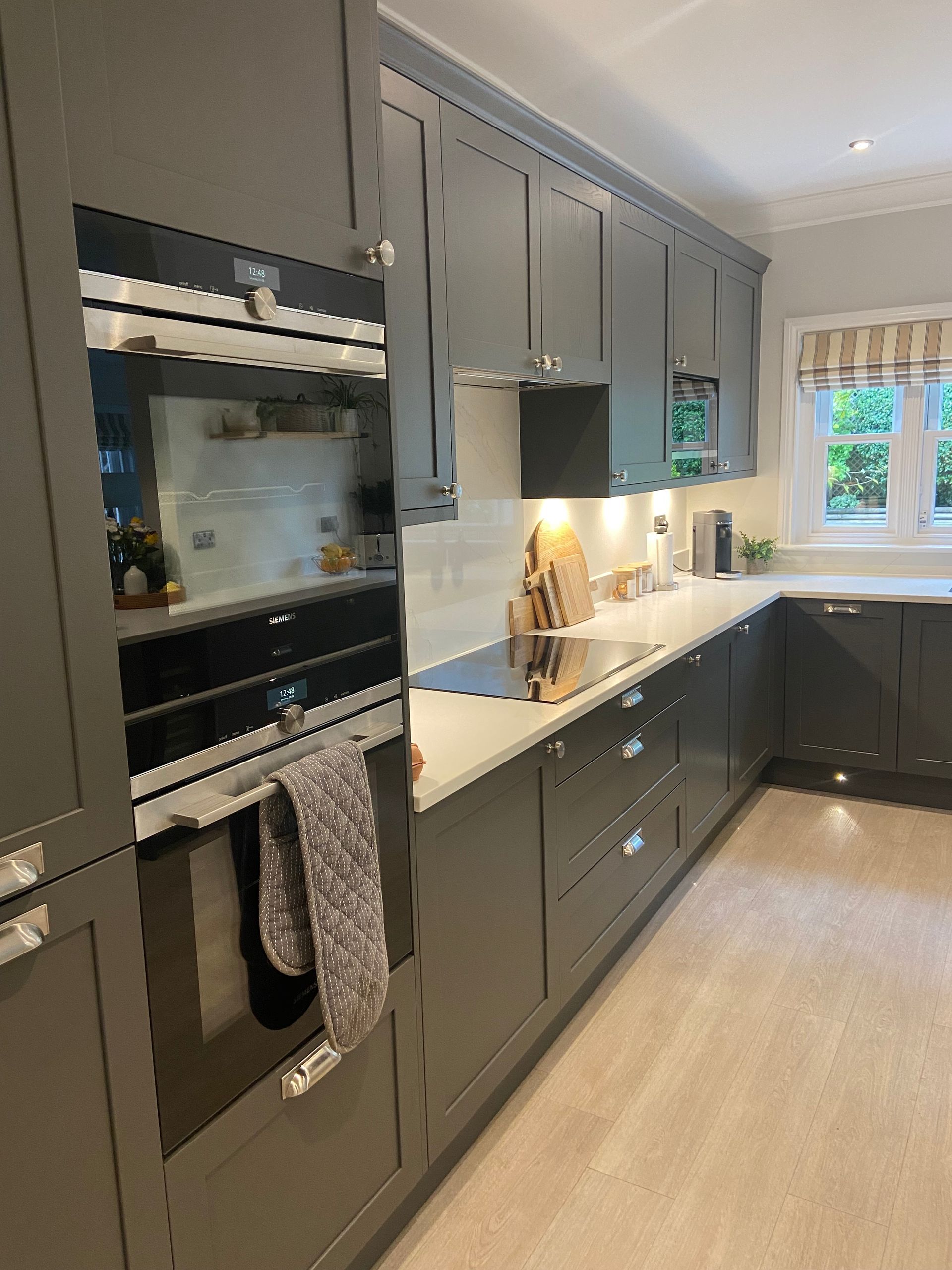 A modern kitchen featuring dark grey cabinets, white countertops, a built-in oven, and a window with striped curtains.